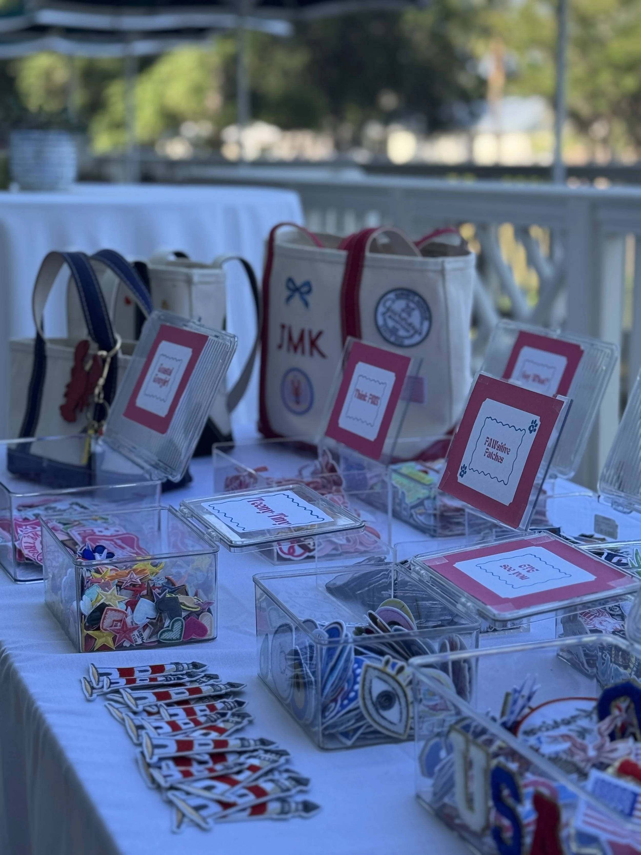 Table with various colorful stickers, badges, and small bags, set up outdoors with a white tablecloth, blurring trees and sky in the background.