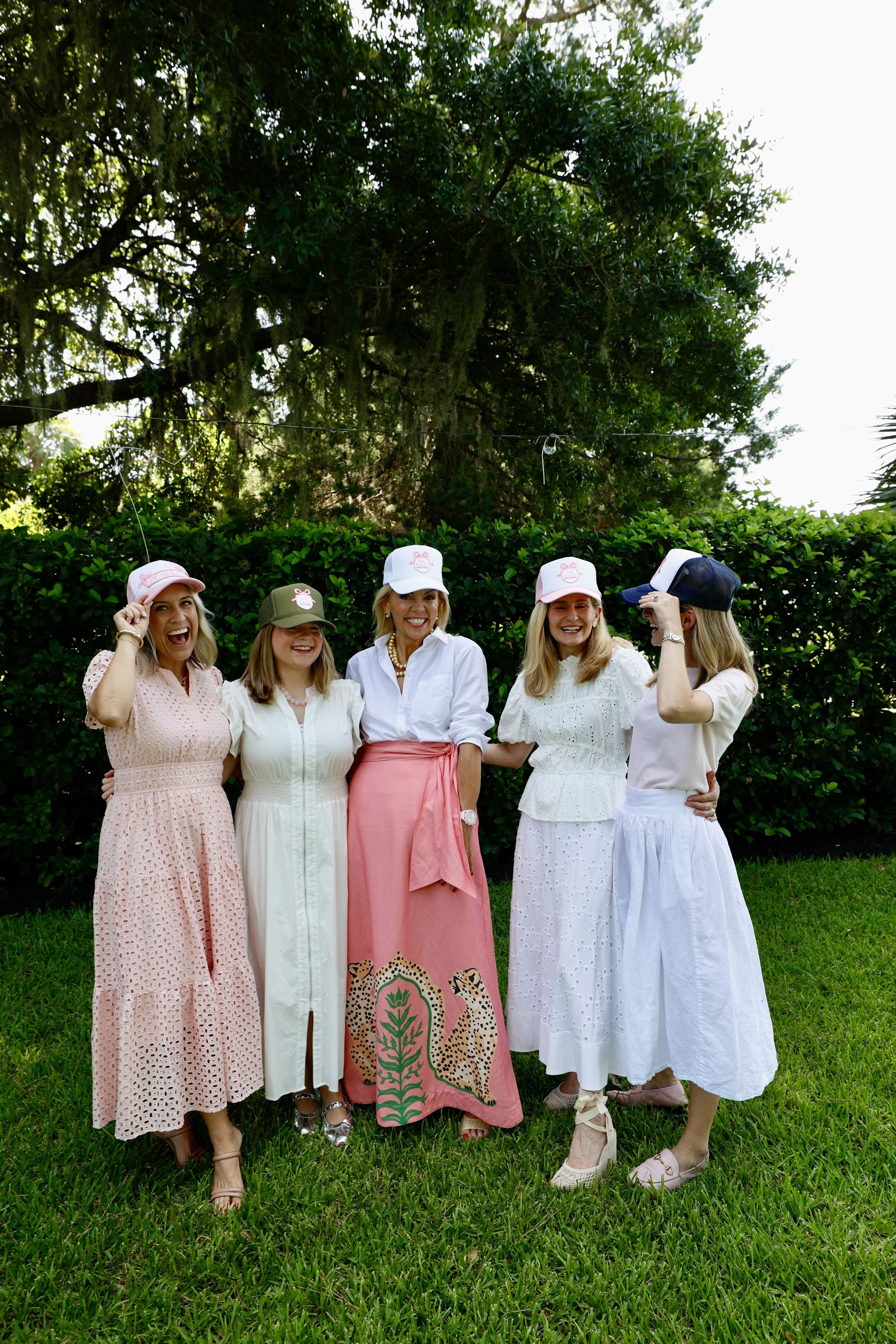 Group of five women, wearing white, pink, and pastel-colored dresses, and caps, standing outdoors on grass with hedge and trees in background, smiling and laughing.