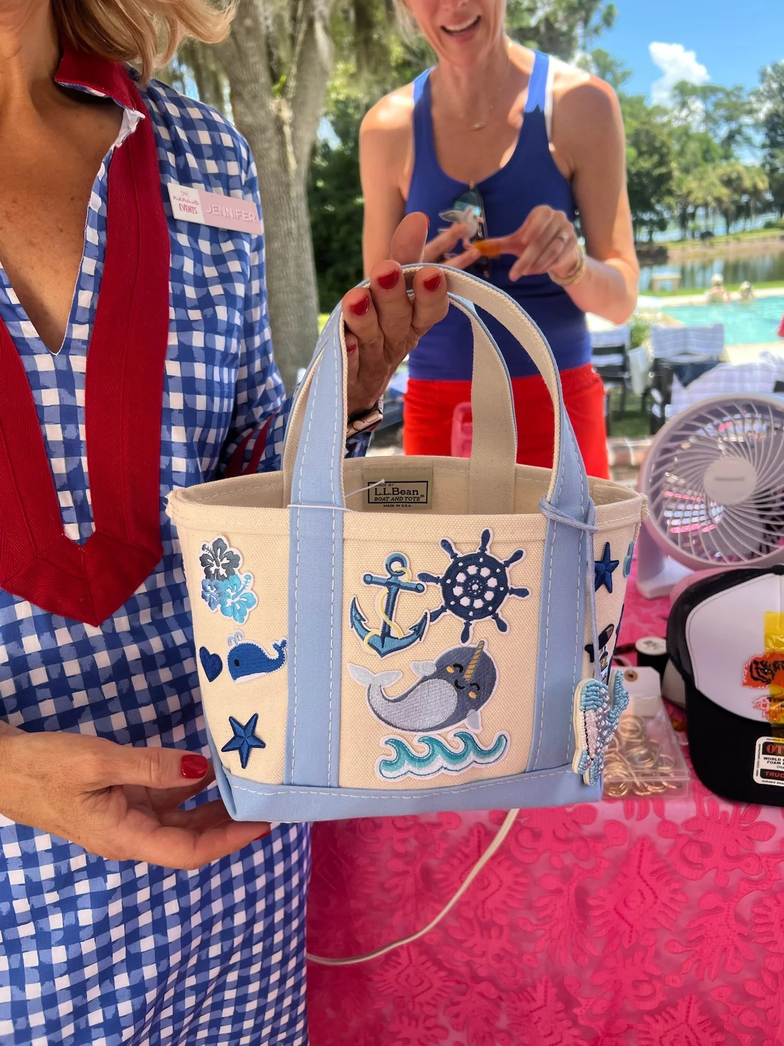 Close-up of two women at an outdoor event during daytime. One woman is wearing a red, white, and blue checkered shirt with a name tag that reads "Jennifer." The other woman is dressed in a blue tank top and red shorts, smiling and pointing at a decor