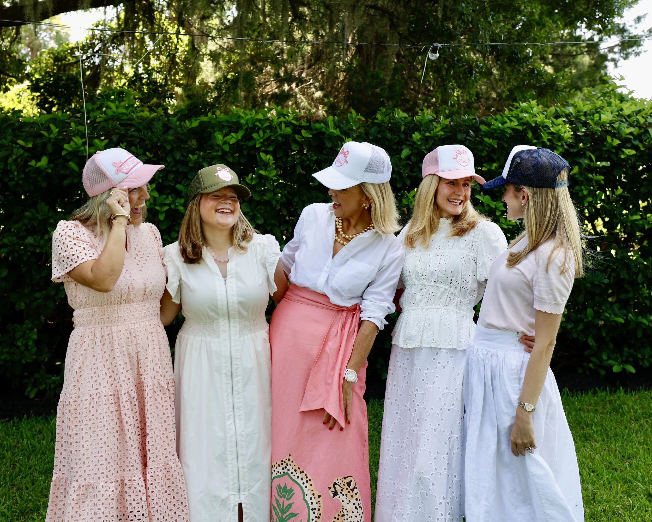 Five women standing outdoors in front of a green hedge, wearing colorful dresses and hats, engaging in conversation and smiling.