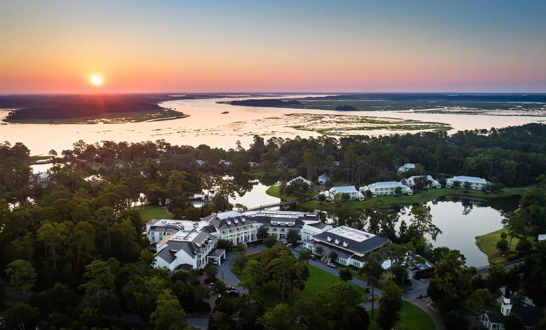 Aerial view of a river winding through a lush green landscape near a residential area during sunset, with colorful sky and calm water reflections.