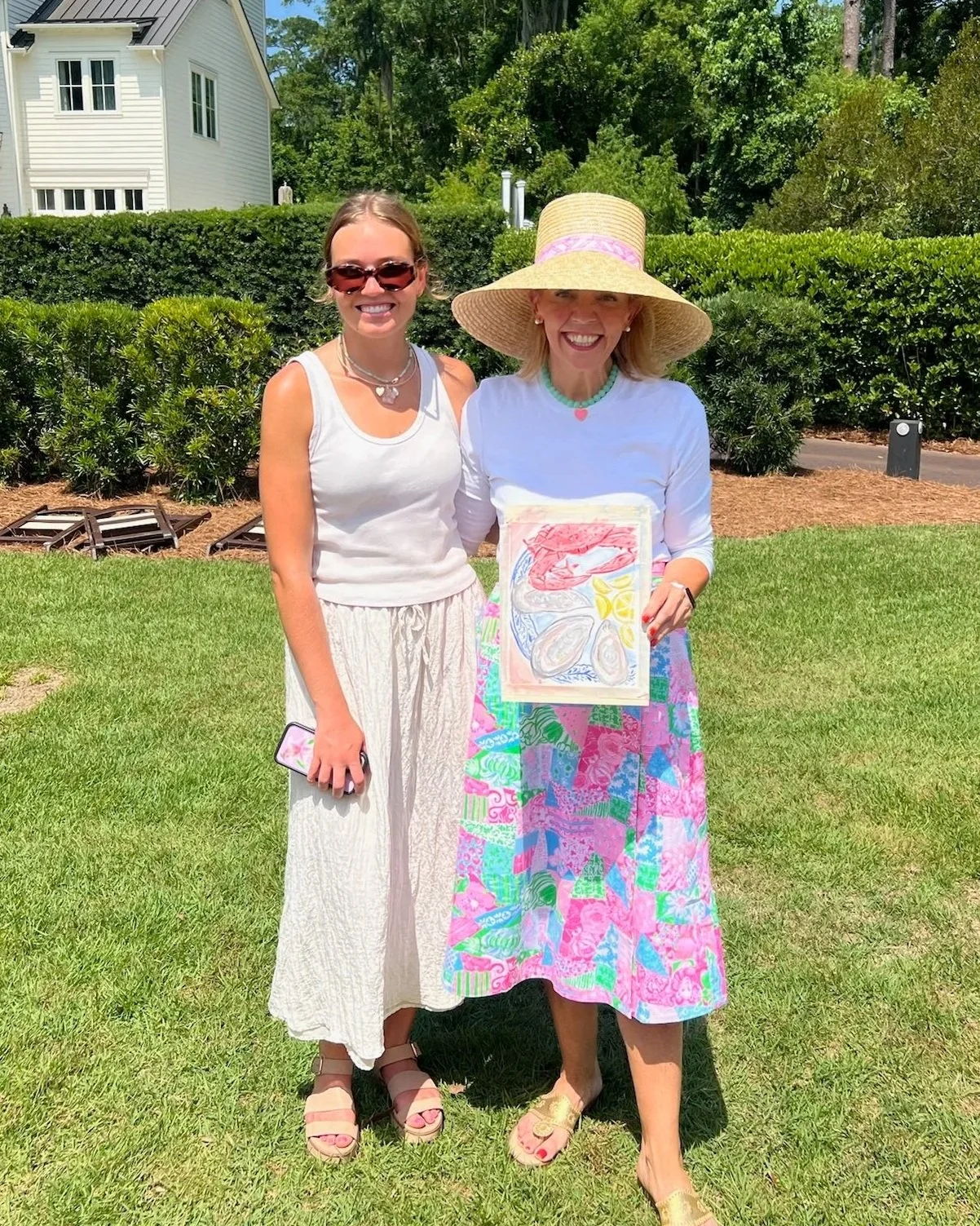 Two women standing outdoors on a lush green lawn holding a colorful drawing of a lobster, shellfish, and lemons.