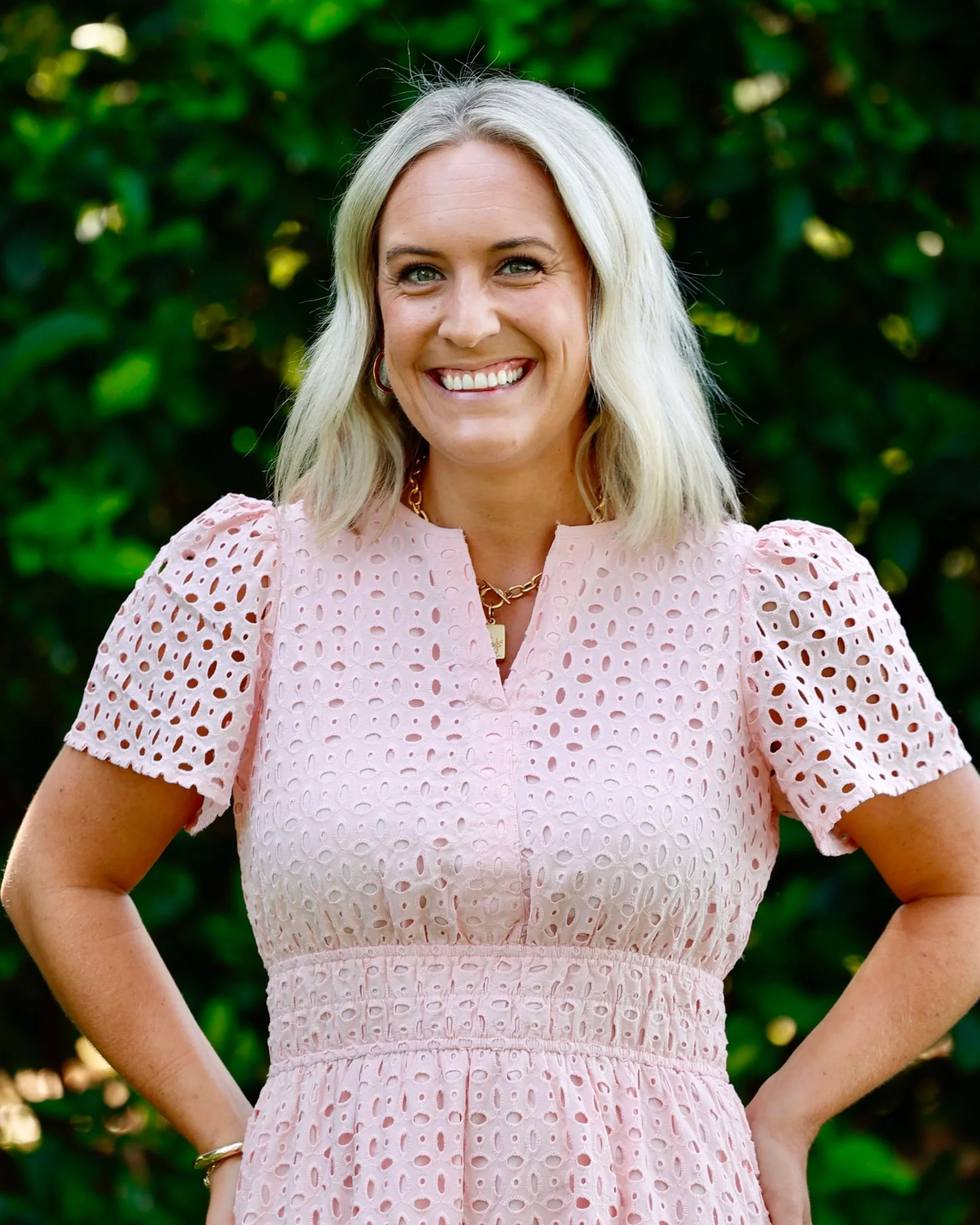 Sam Reddy smiling and wearing a pink lace dress, standing outdoors with green foliage in the background.