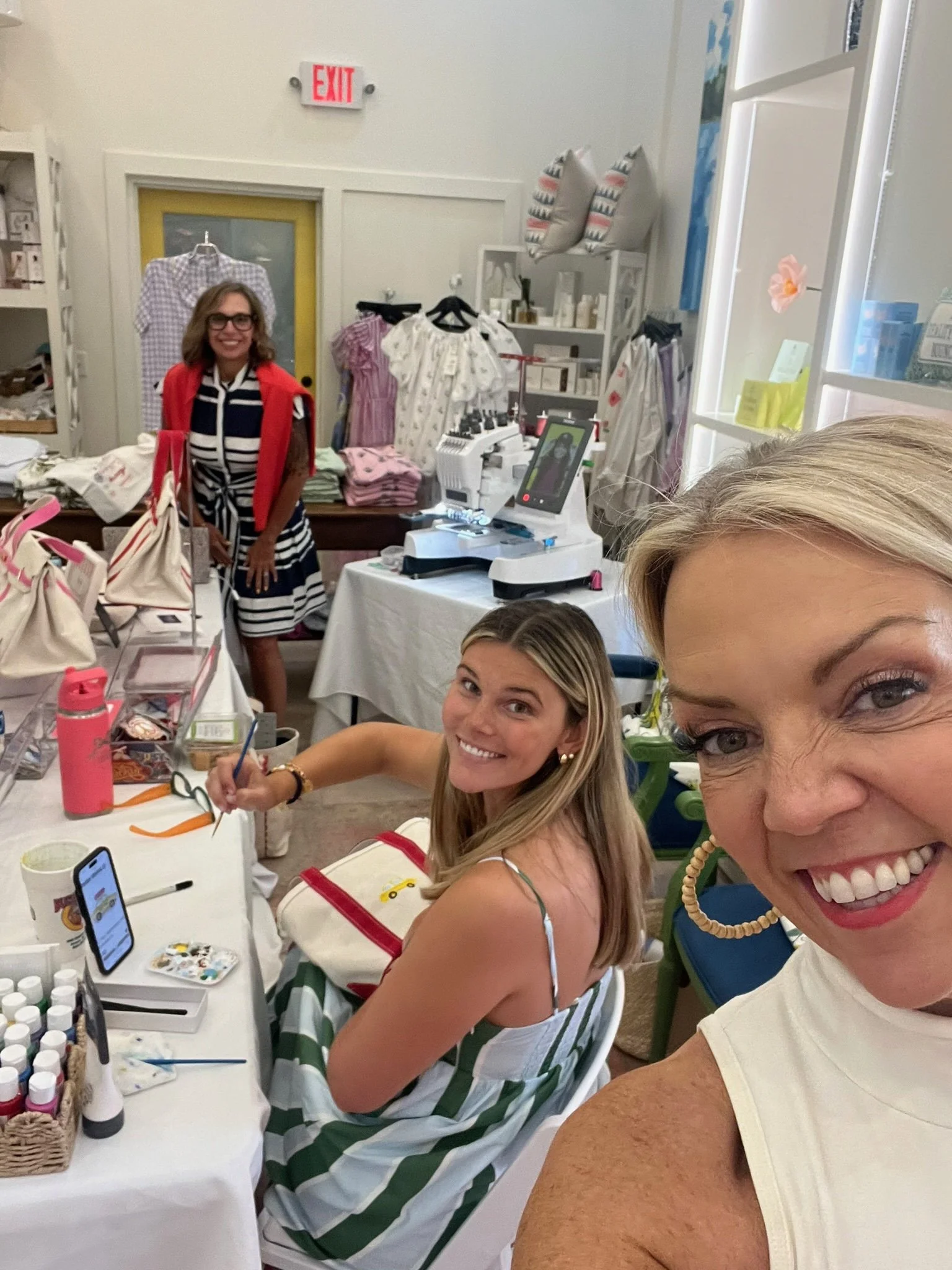 Smiling women at a crafting or art station with paints, brushes, and craft supplies, surrounded by clothing and decorative pillows in a well-lit room.