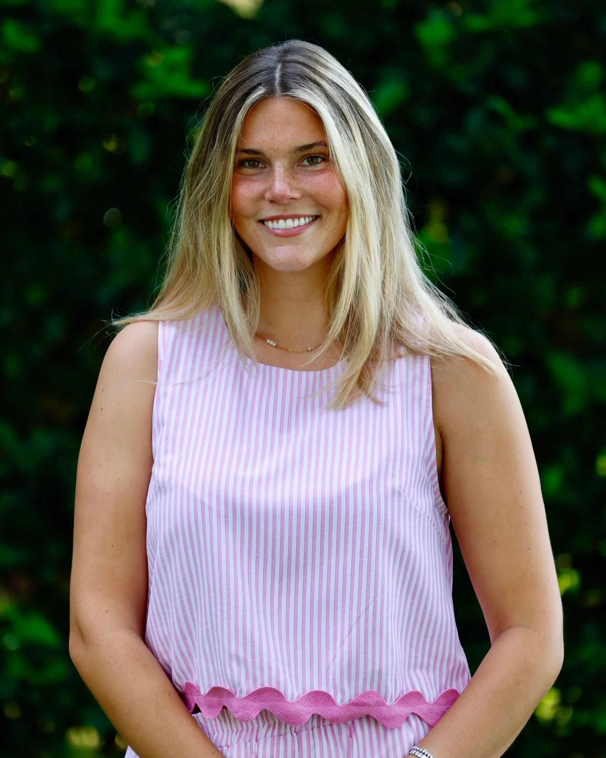 Megan Bast wearing a pink and white striped dress with ruffle detail near the hem, standing outdoors with greenery in the background.