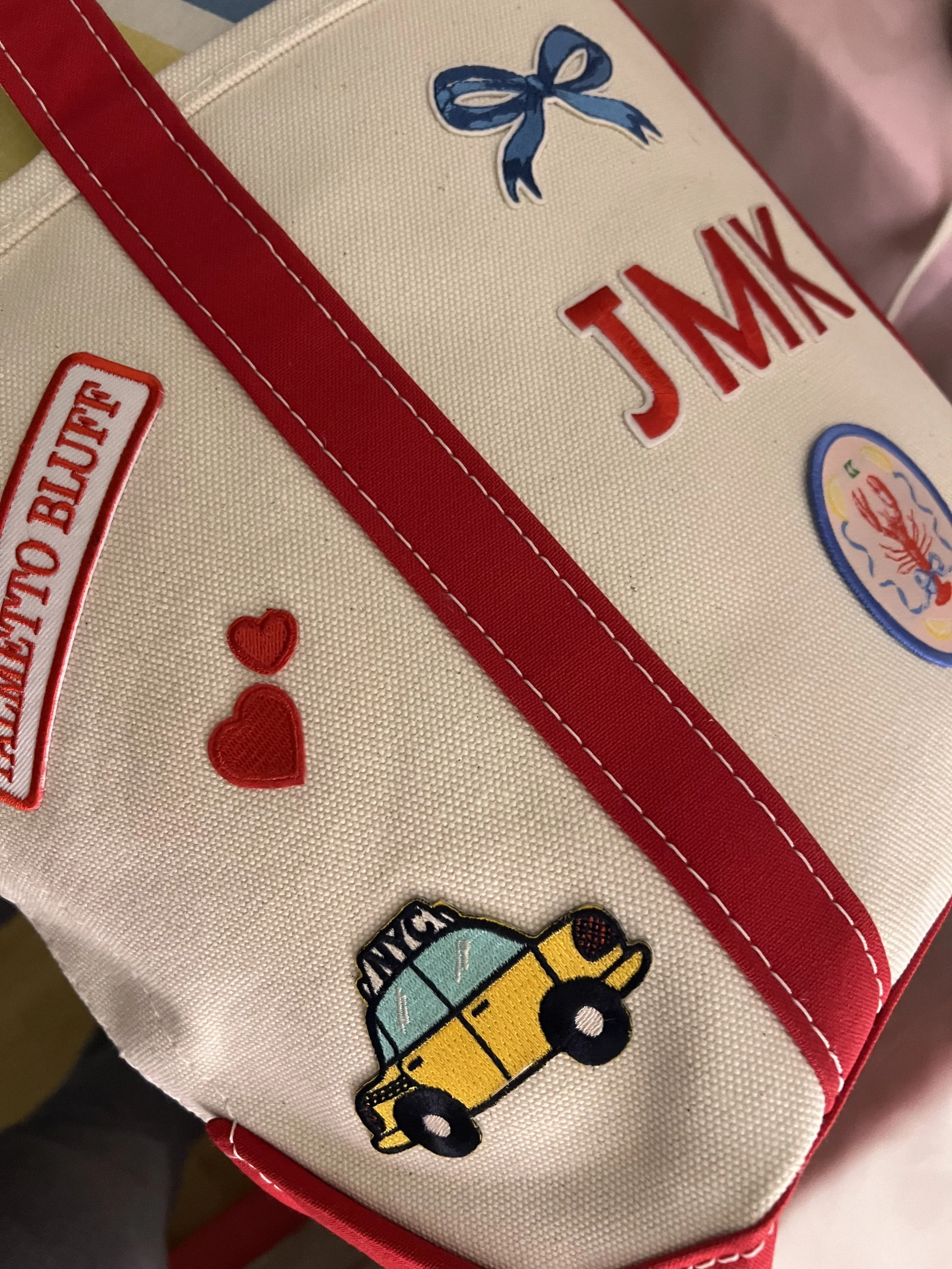 L.L. Bean Boat and Tote decorated with several patches, including a patch of a yellow school bus, a blue bow, a heart, and a lobster. The backpack is mainly beige with red straps and trim.