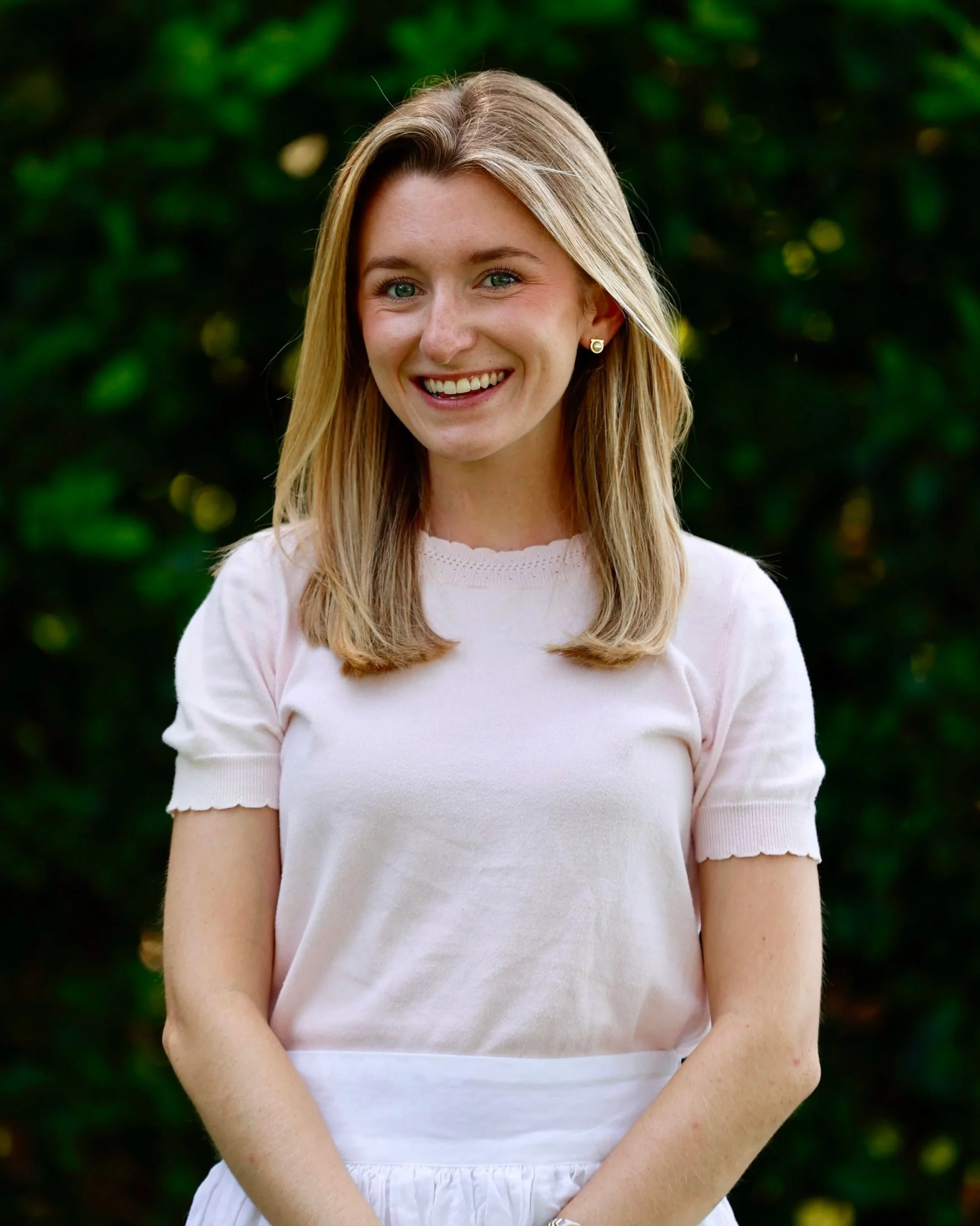 A young woman with long blonde hair, smiling, wearing a pale pink short-sleeved top and earrings, standing outdoors against a backdrop of green foliage.