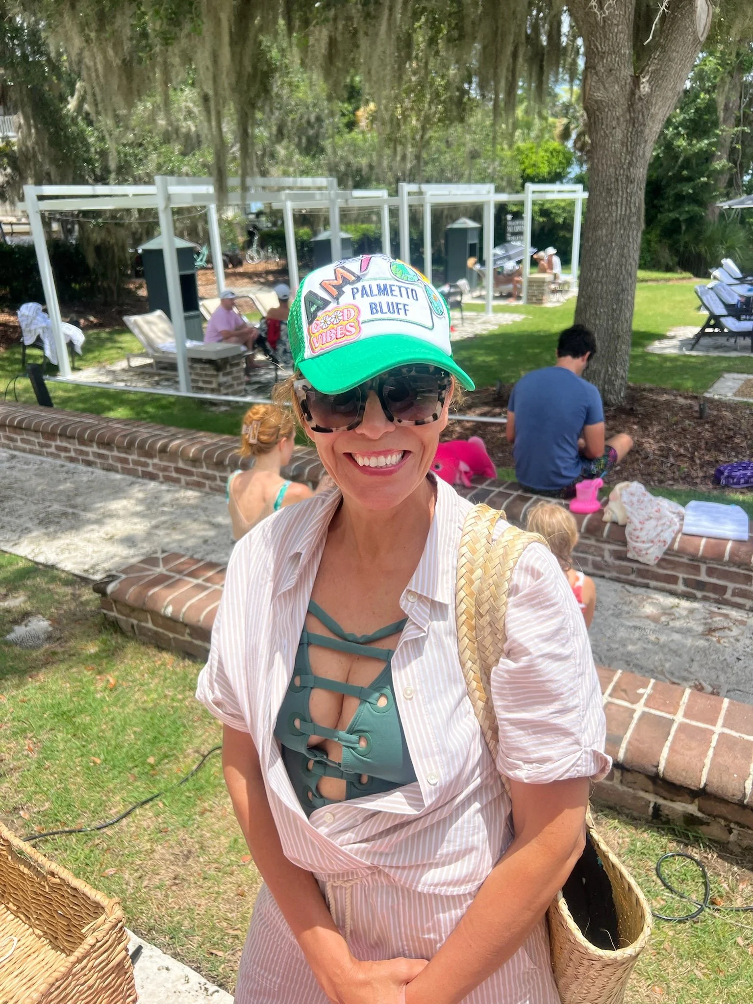A woman smiling at the camera, wearing sunglasses, a cap that says "Palmetto Bluff Vibes," a striped shirt, and a swimsuit top, standing outdoors in a park-like setting with trees, benches, and other people in the background.