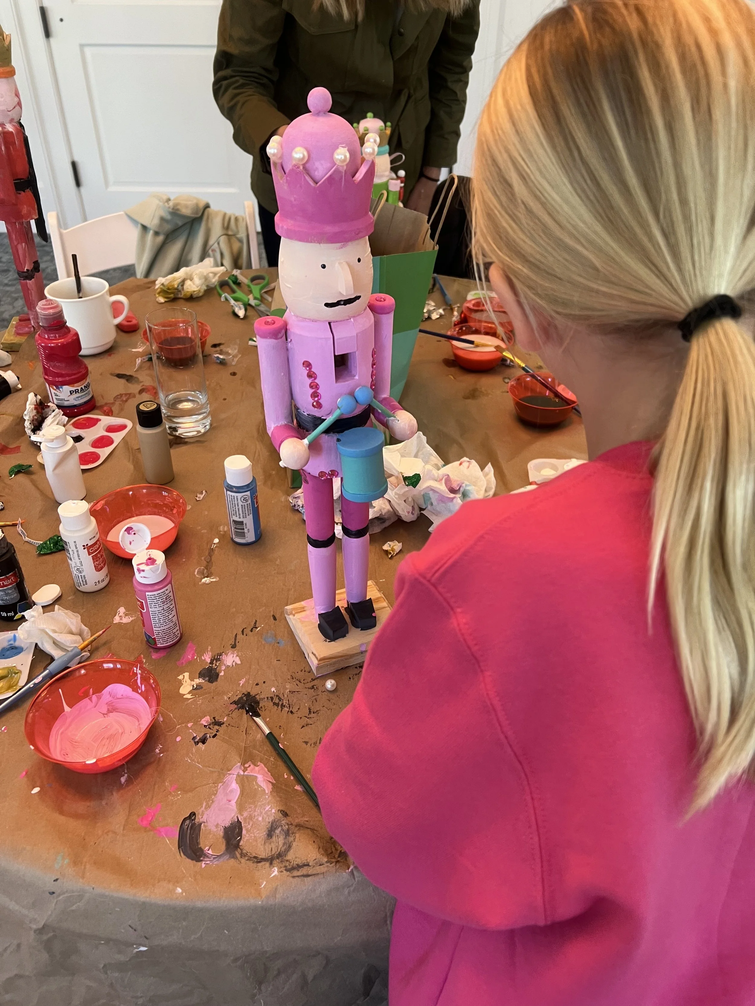 People creating a craft project with a large painted wooden nutcracker figure topped with a pink crown and decorated with pink and blue accessories, surrounded by art supplies and paint on a table.