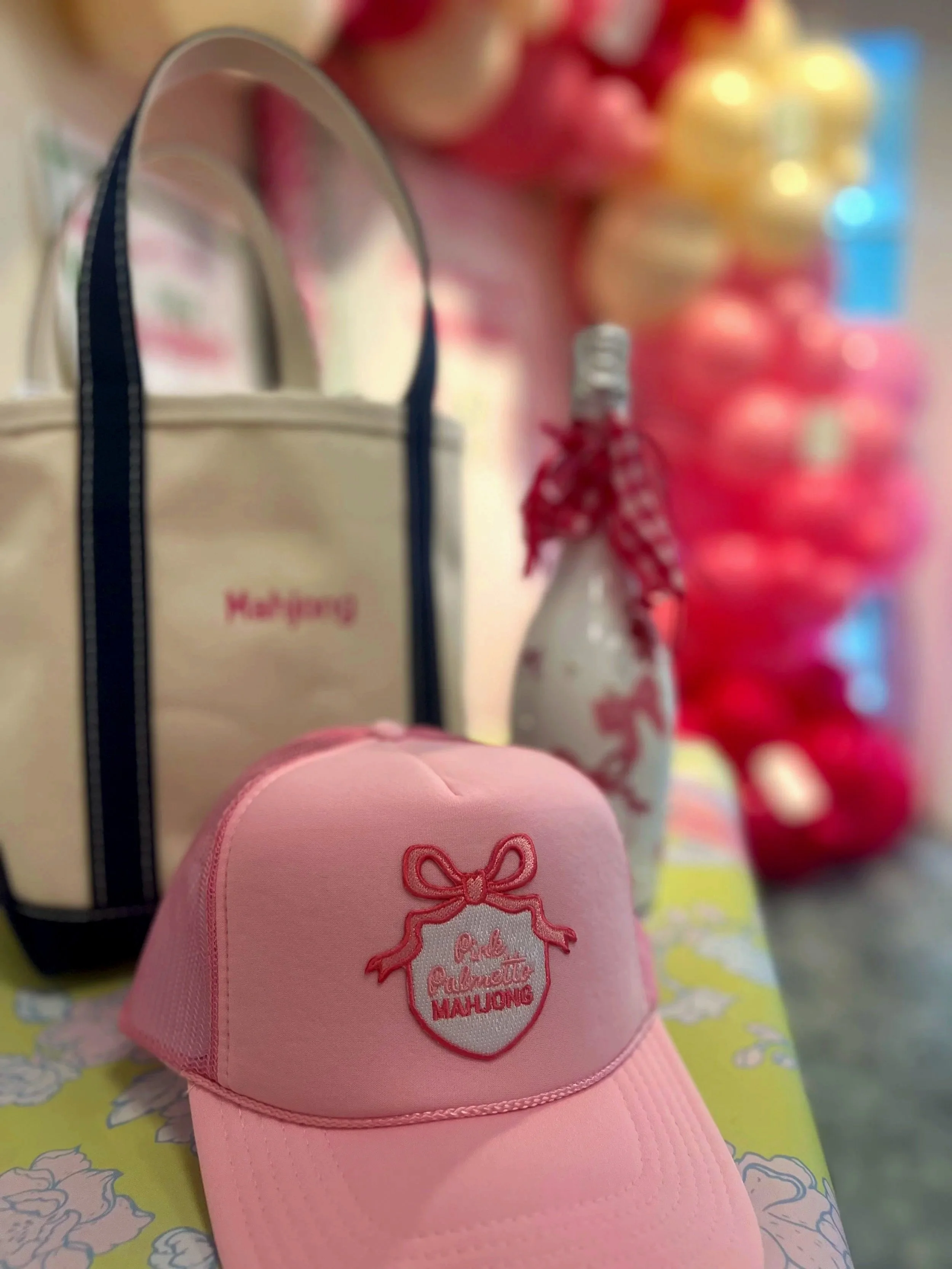 Close-up of a pink cap with embroidered text and a bow design, in front of blurred colorful objects in the background.