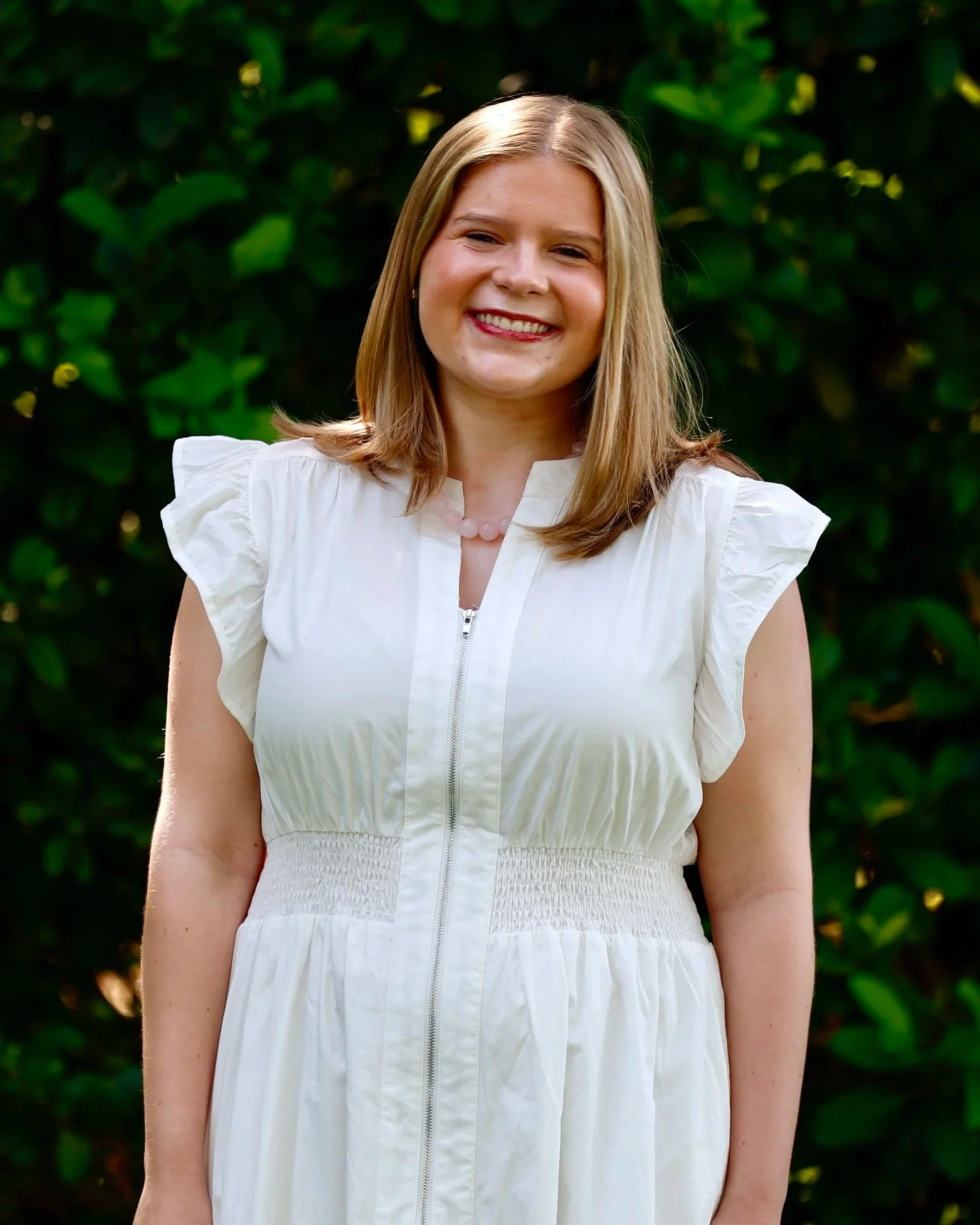 Landrum Barnes wearing a white dress with a zipper, standing outdoors against a background of green foliage, smiling.