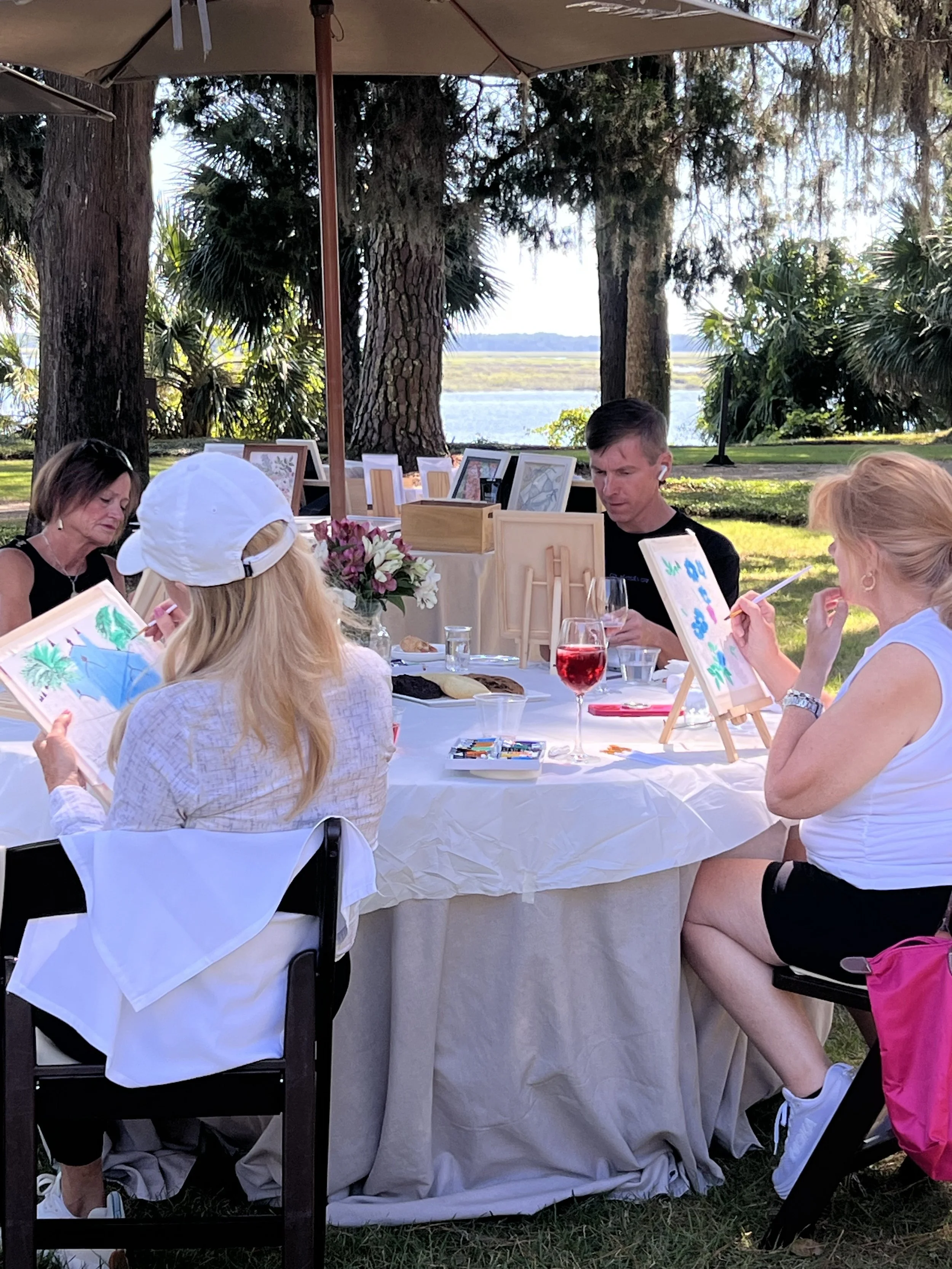 People sitting around a table outdoors painting pictures, with trees and a body of water in the background.