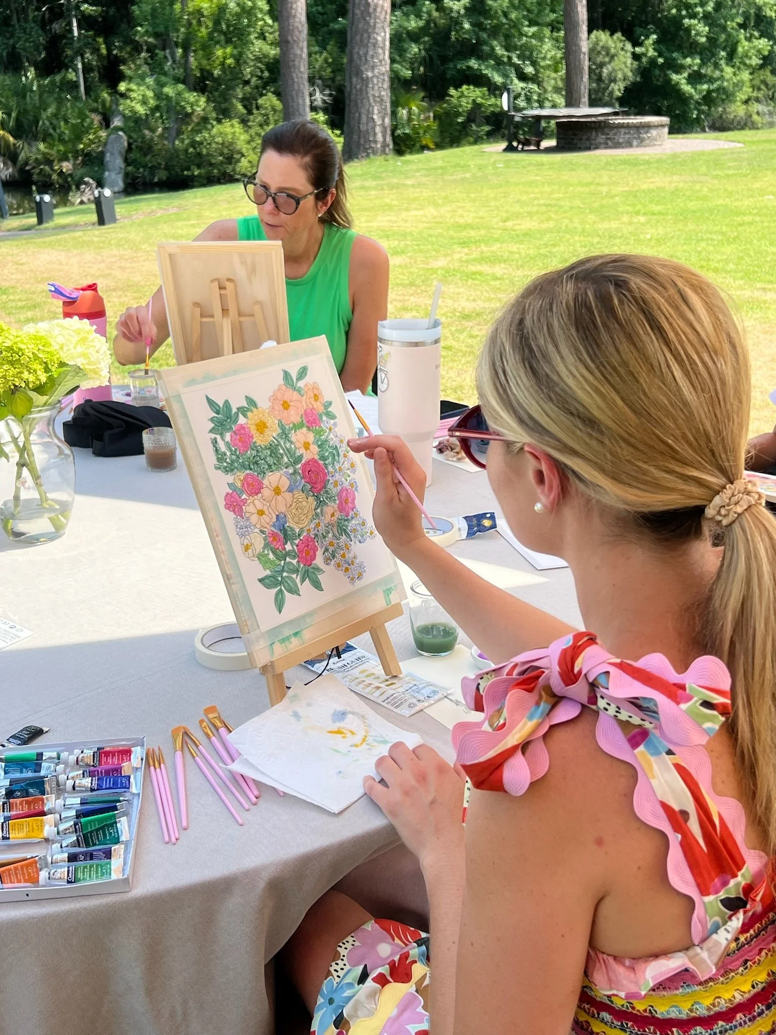 Two women painting floral art outdoors at a table with art supplies, surrounded by greenery and trees.