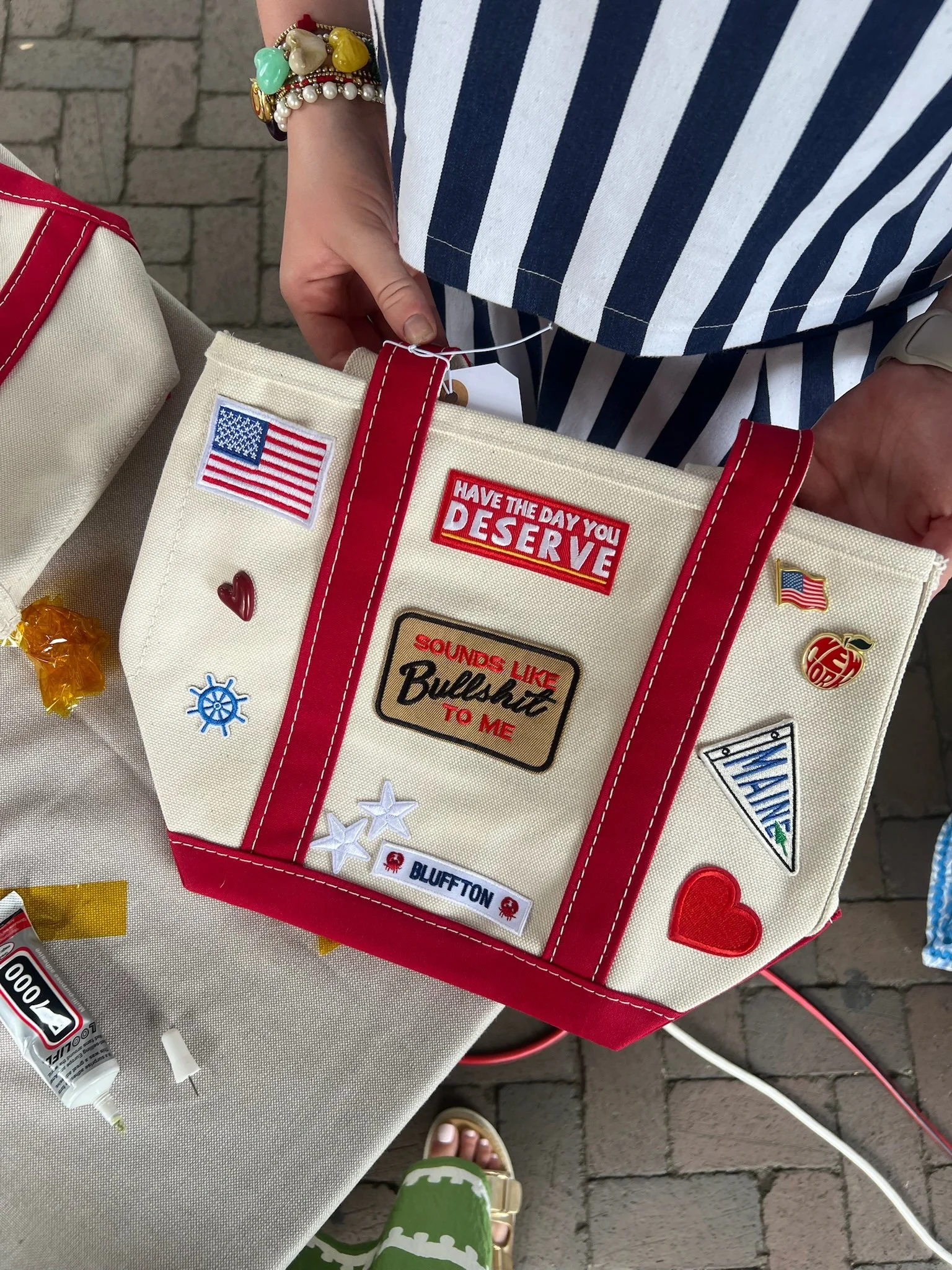 Close-up of a woman holding a white tote bag decorated with patches and pins, including an American flag and a red heart, against a brick pavement background. The woman is wearing a blue and white striped shirt, and her accessories include colorful b