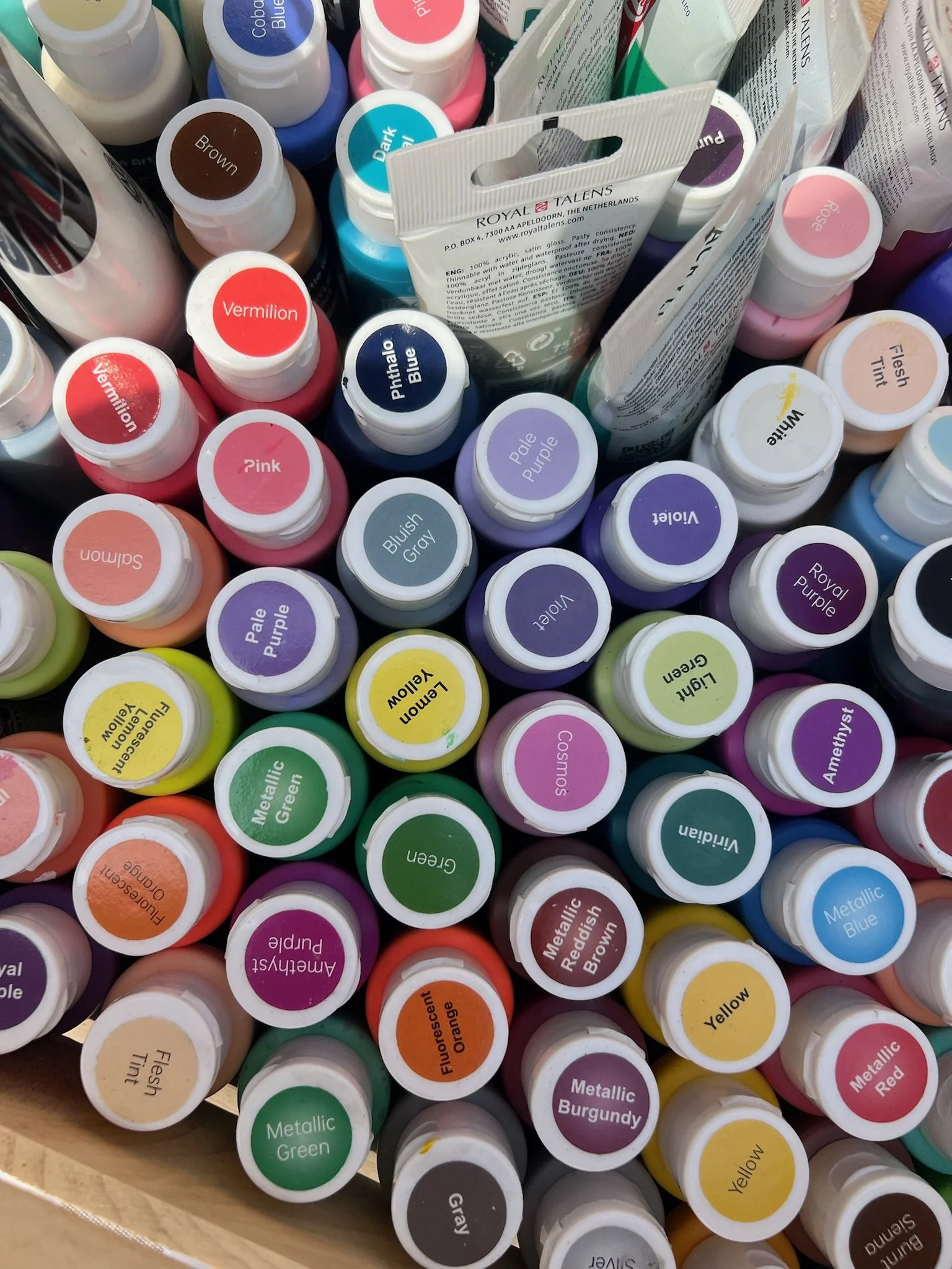 A collection of acrylic paint bottles with various color labels, including brown, dark teal, pink, violet, cream, yellow, green, and black, arranged in a container.