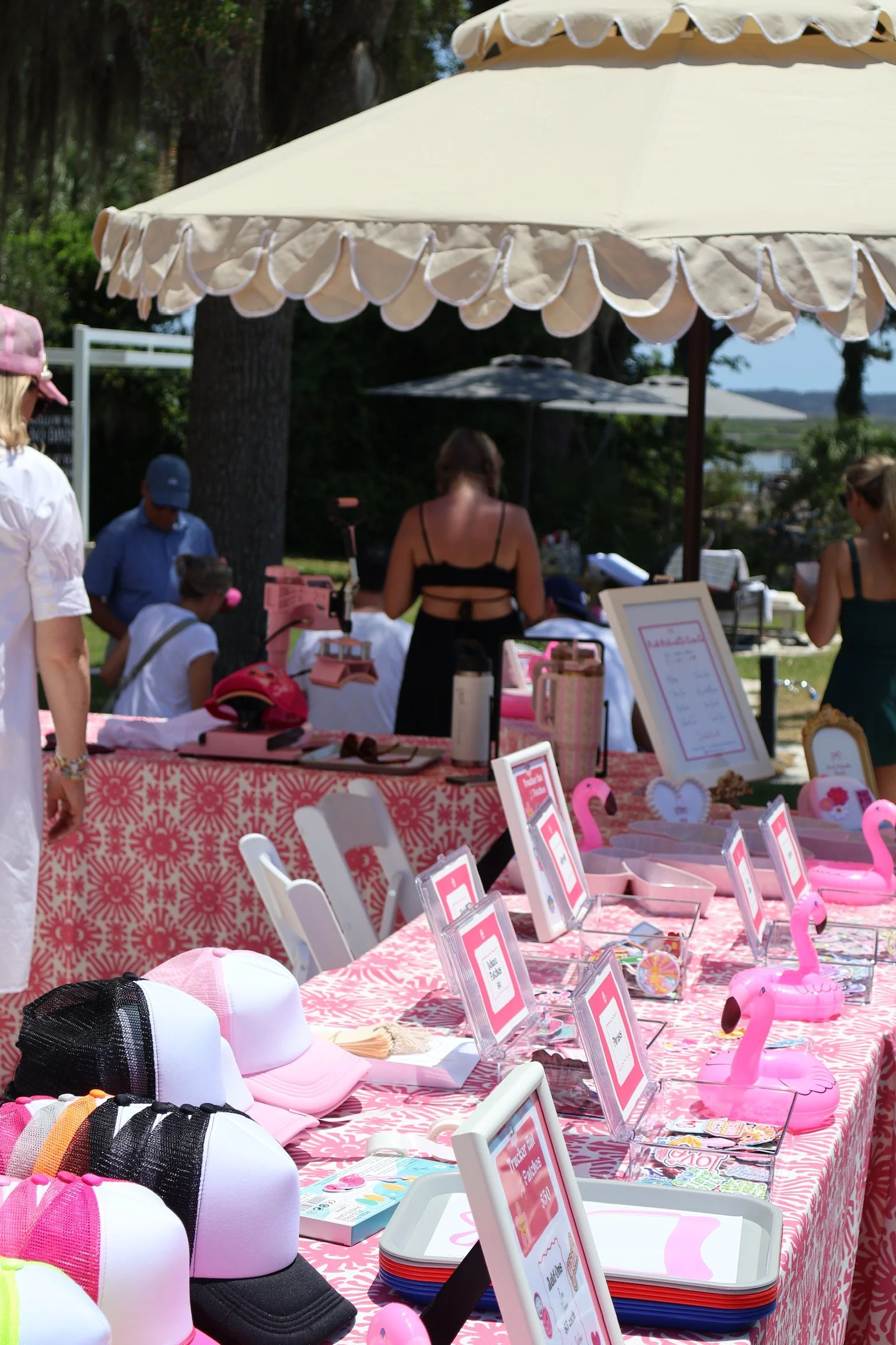 A colorful outdoor patch bar pink-themed items, pink flamingo decorations, hats, and display signs on a pink patterned tablecloth under large umbrellas.