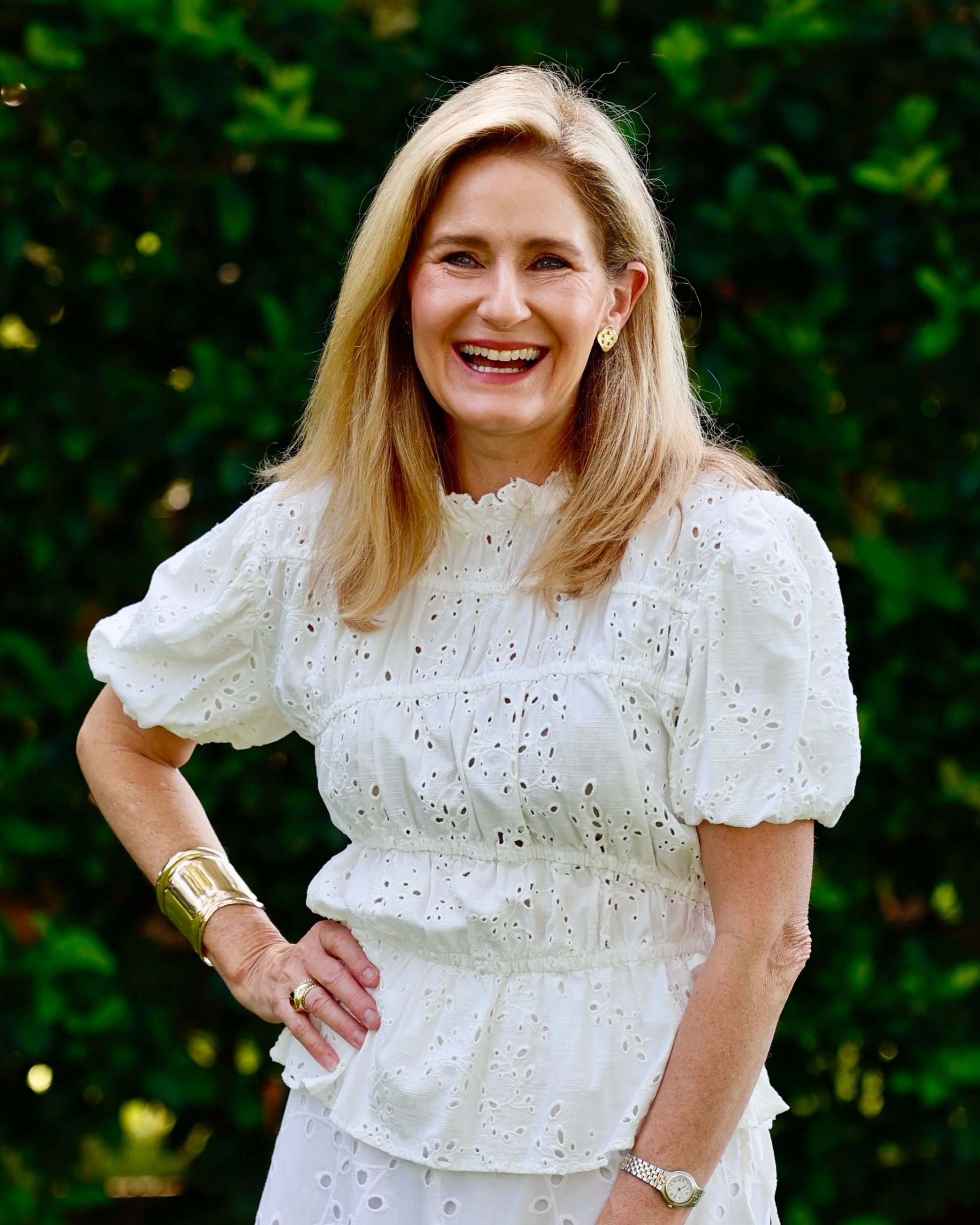 Carrie Collins wearing a white eyelet dress with puffy sleeves, smiling with her left hand on her hip, surrounded by green foliage.
