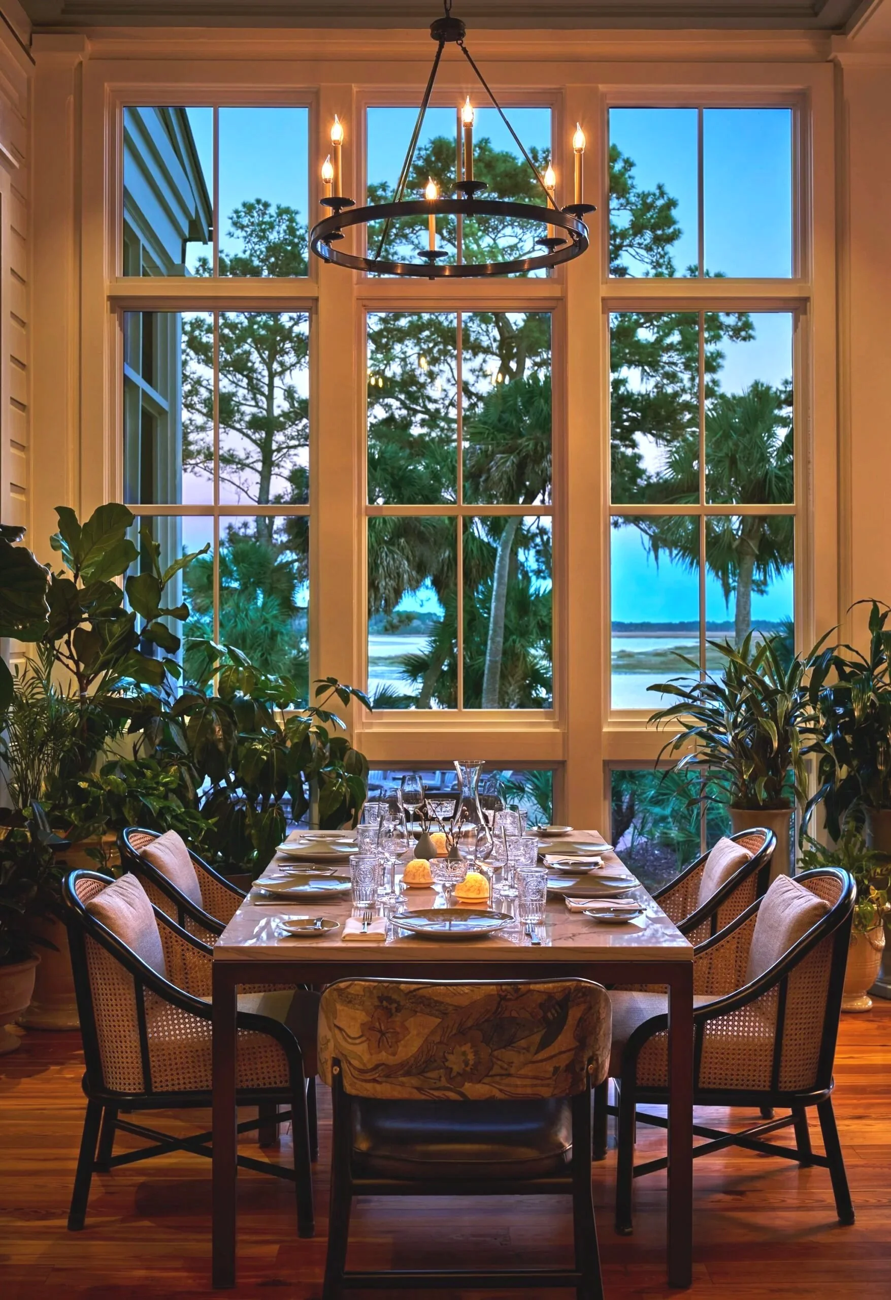 Elegant indoor dining room with a large window view of trees and water, decorated with plants, a chandelier, and a set dining table.