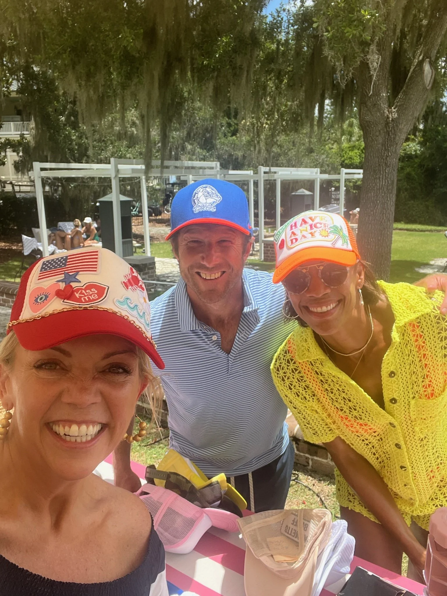 Three smiling women and one man taking a selfie outdoors on a sunny day, with trees and a porch in the background. They are wearing colorful hats and casual summer clothing.