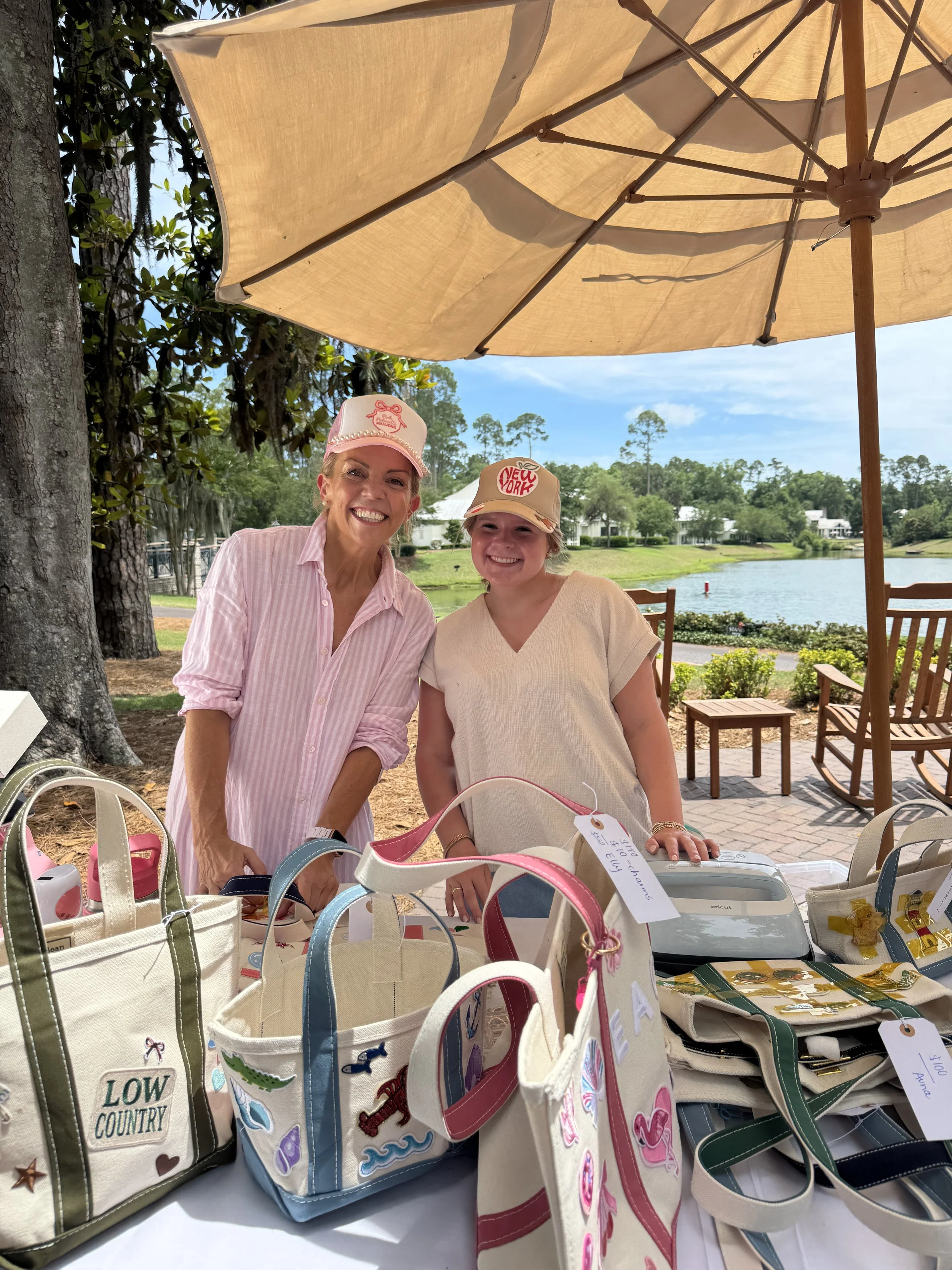 Two women smiling under a large beige outdoor umbrella with a lake and trees in the background. The table in front of them displays various colorful embroidered tote bags for sale.