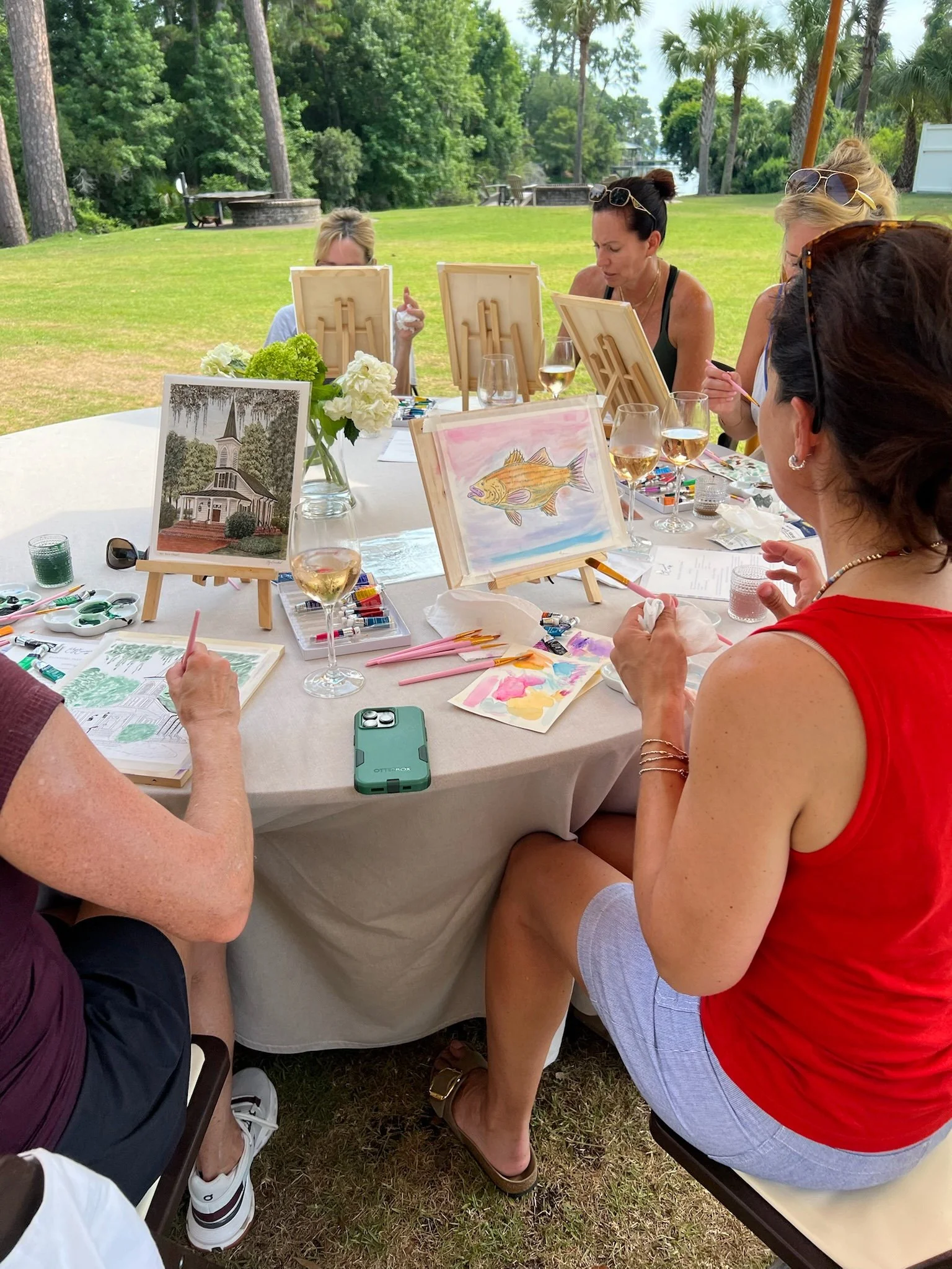 A group of women painting outdoors at a long table with art supplies, framed pictures, and glasses of wine, in a lush green park with trees.