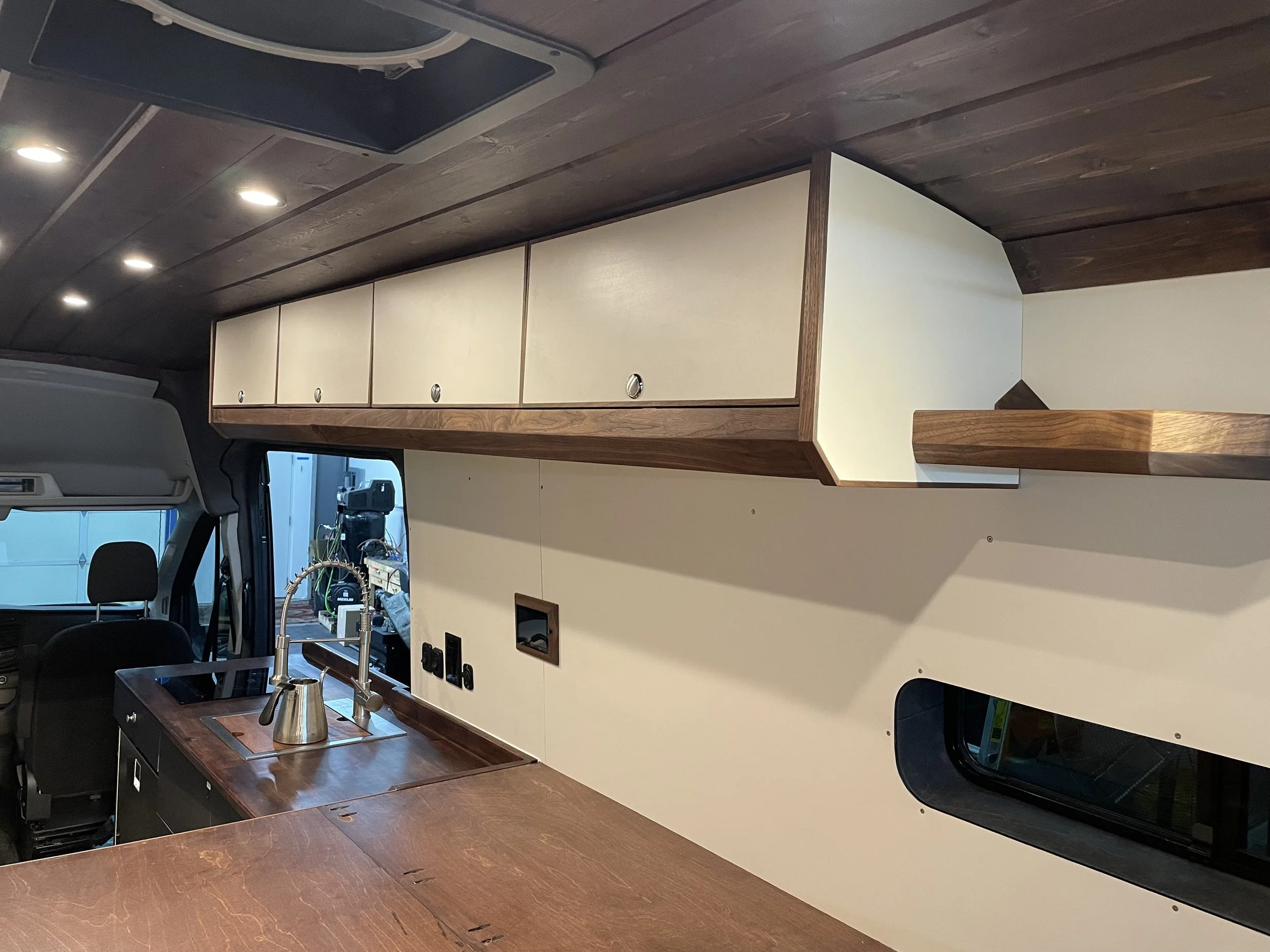 Interior of a camper van with a wooden countertop, a sink with a faucet, white cabinets above, and a small window.