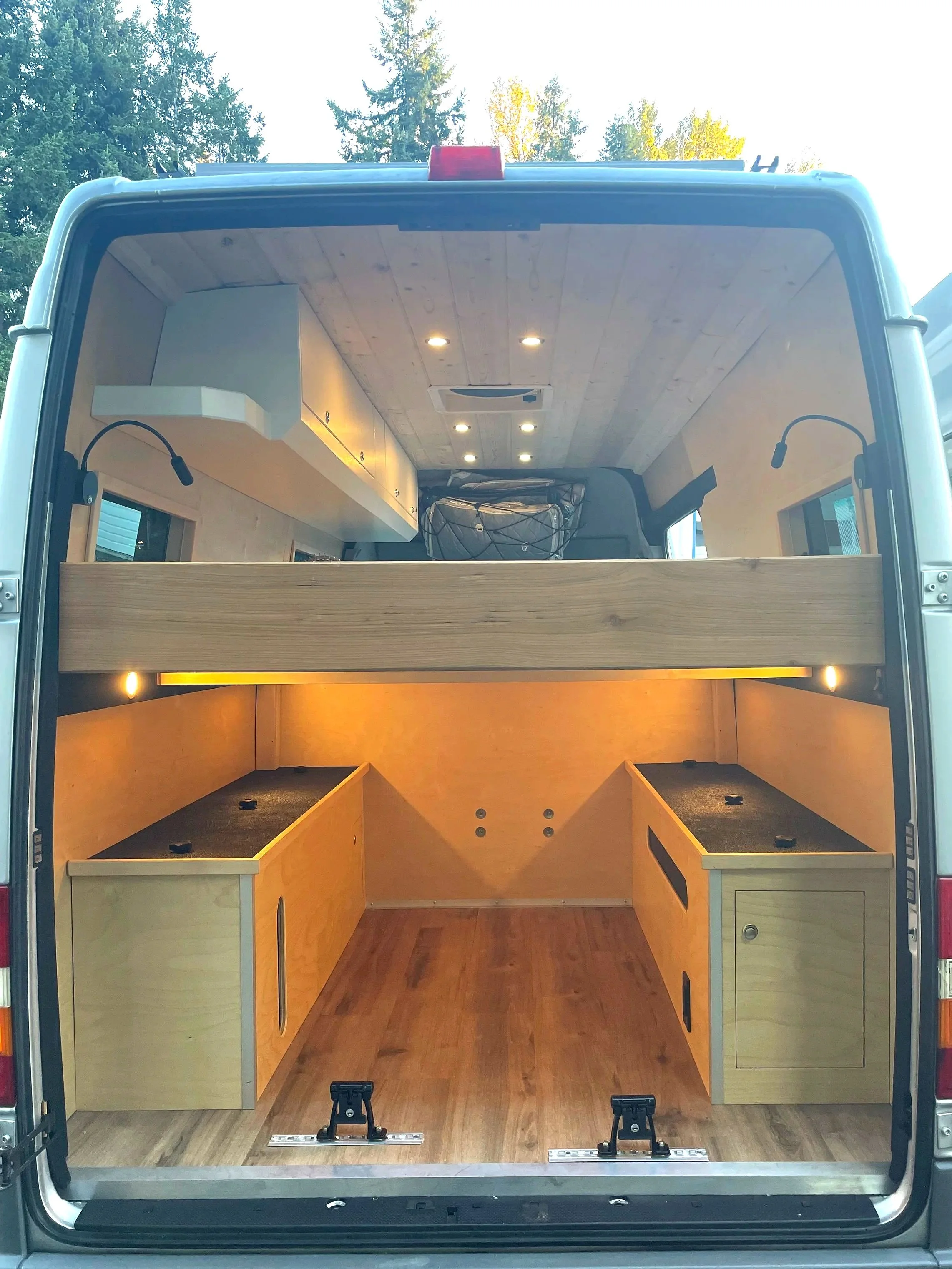 The interior of a converted camper van with wooden cabinetry and flooring, LED lights, and a loft bed area.