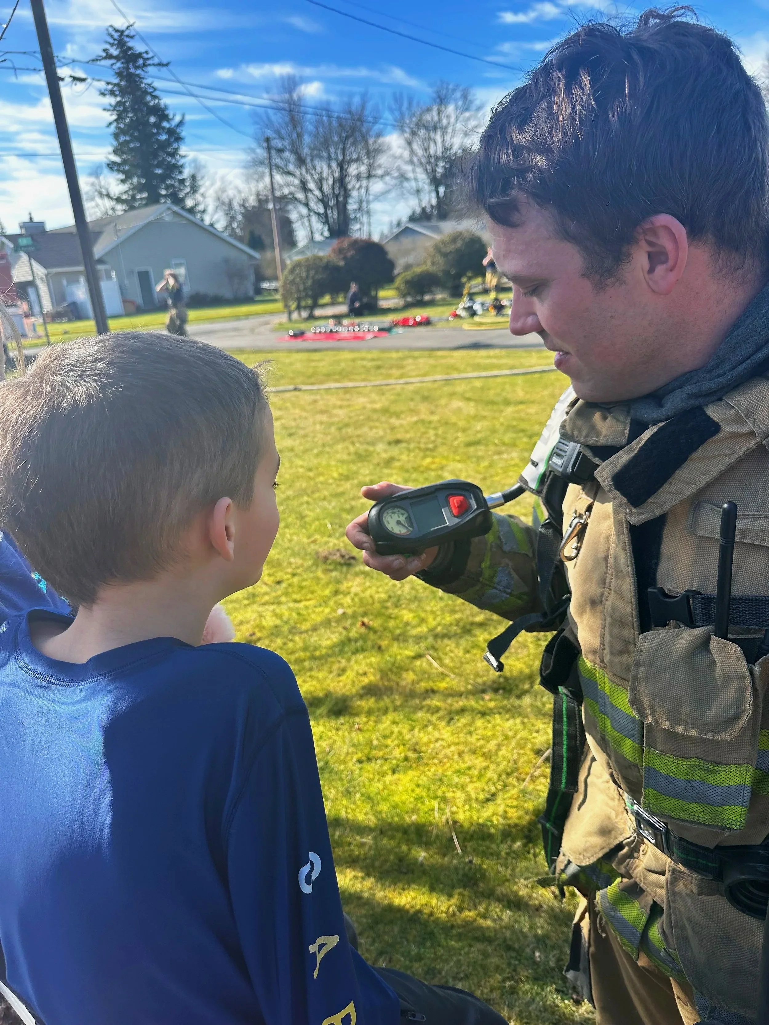 A firefighter showing a young boy a handheld device, standing outdoors on a sunny day with houses and trees in the background.