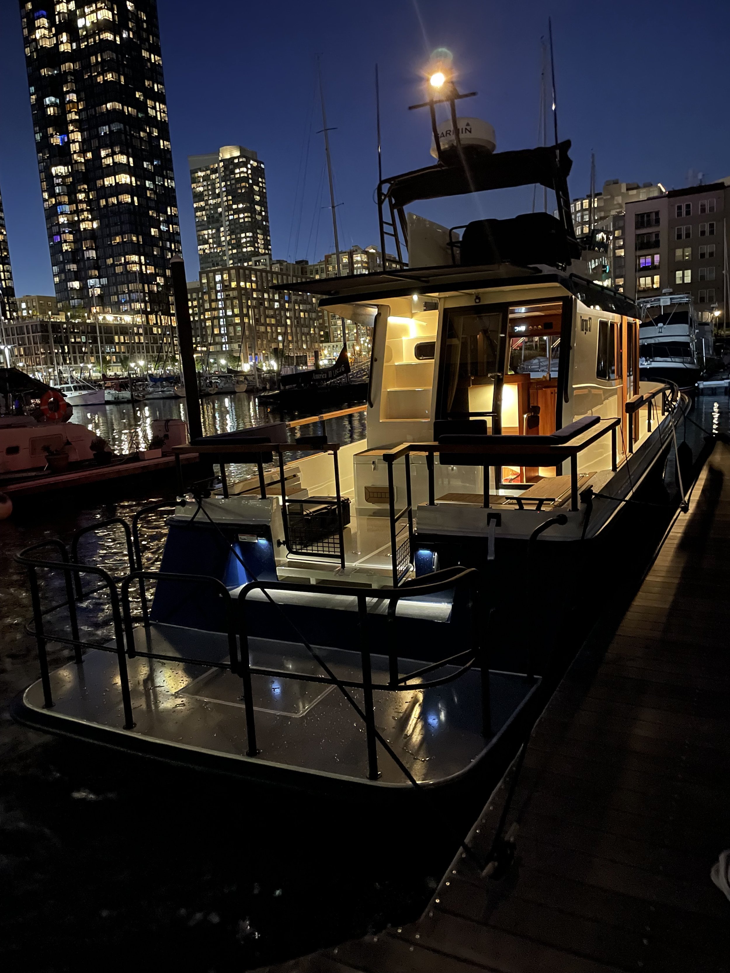 Nighttime scene at a marina with a houseboat docked at a wooden pier, city lights and high-rise buildings in the background.
