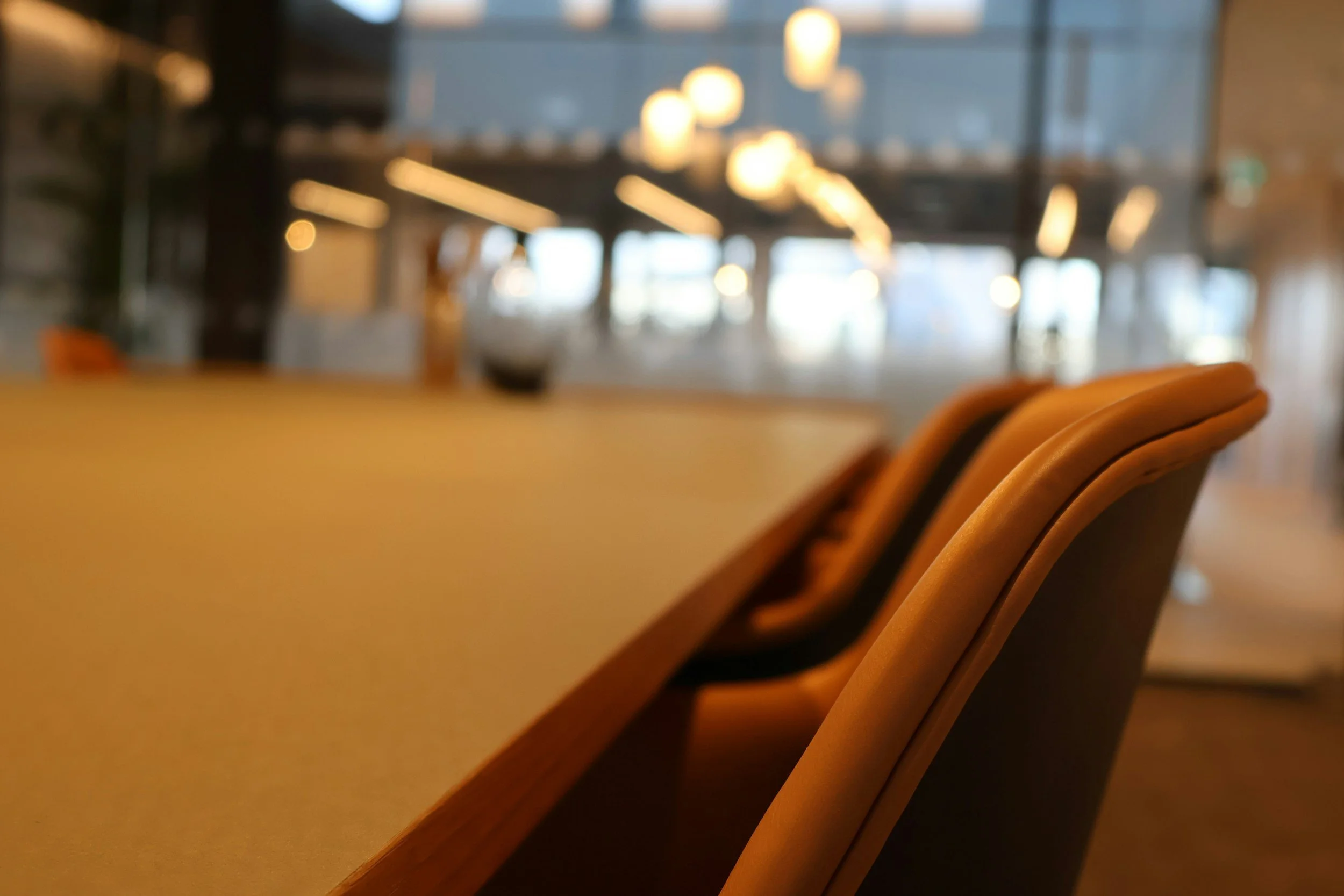A blurry image of a conference room or dining area with a long wooden table and beige cushioned chairs, with warm lighting and large windows in the background.