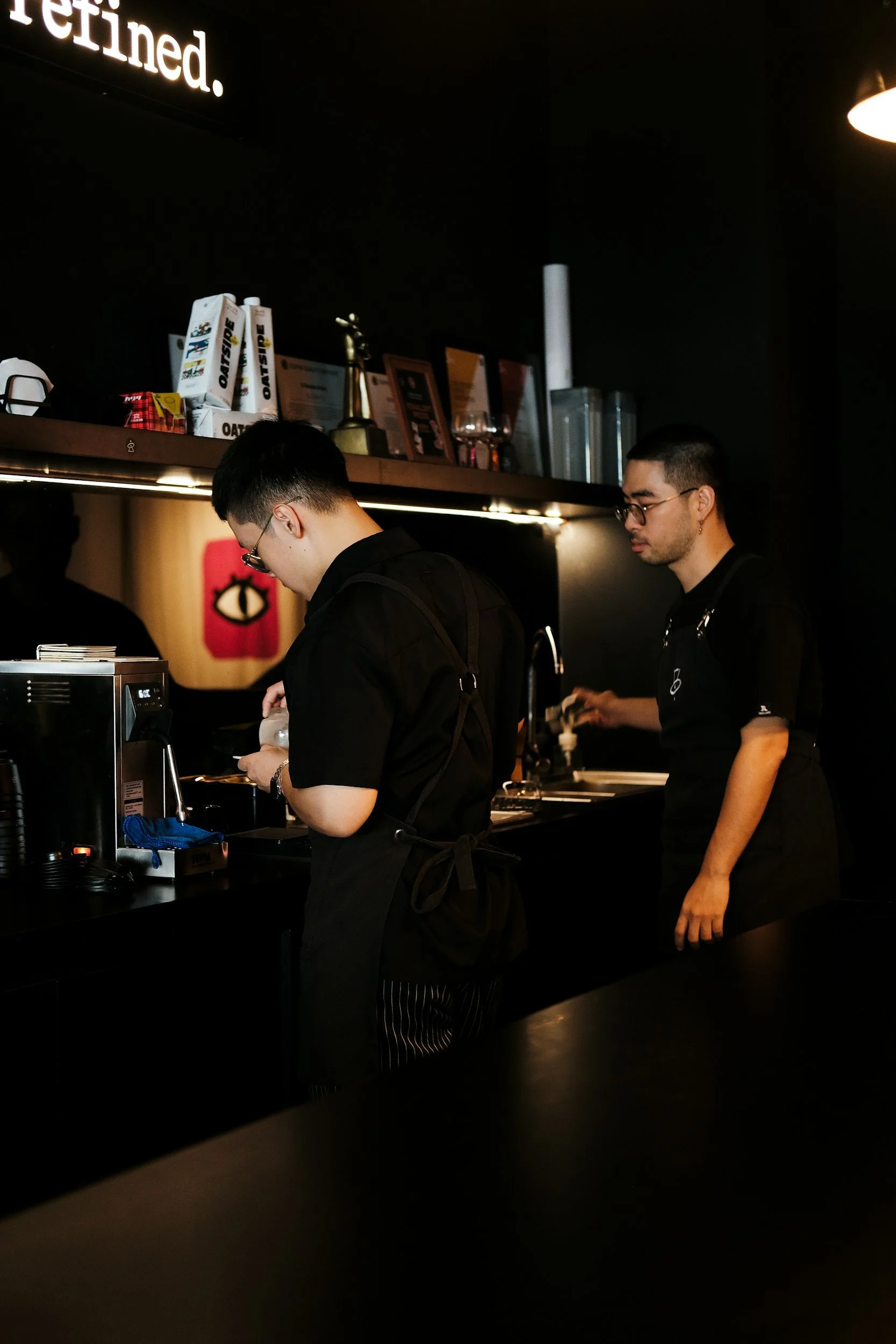 Two male baristas working behind the counter in a dark, modern coffee shop, wearing black shirts and aprons, with coffee-making equipment and framed certificates or pictures on the wall behind them.