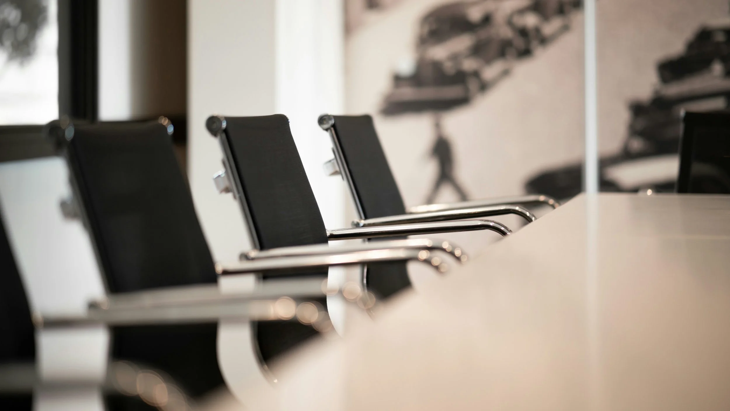 Empty black conference chairs with chrome armrests arranged around a white conference table in a meeting room, with a blurred wall art of a person walking in the background.  Ready for job interviews