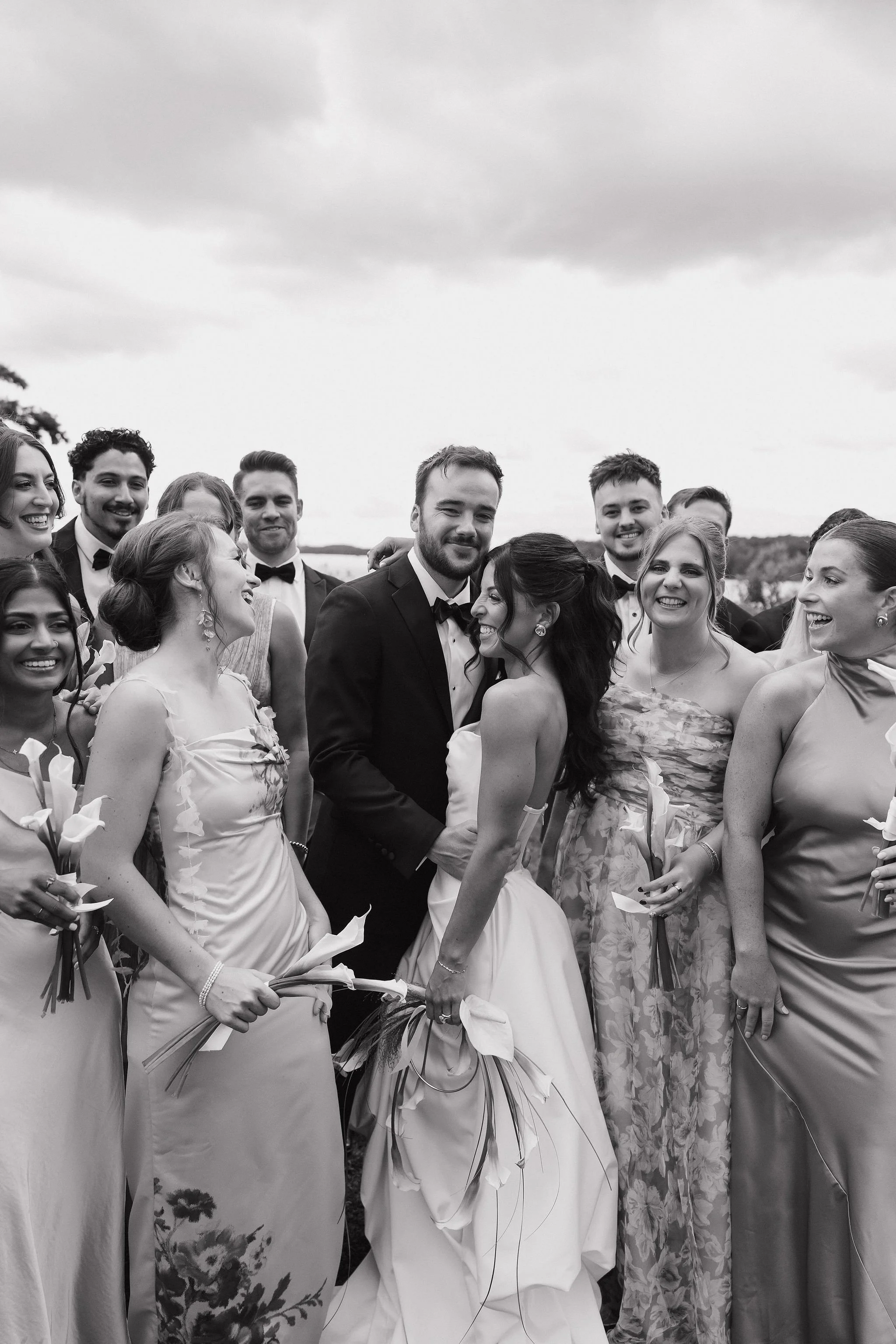 Black and white photo of a bride and groom surrounded by bridesmaids and groomsmen outdoors, celebrating at a wedding.