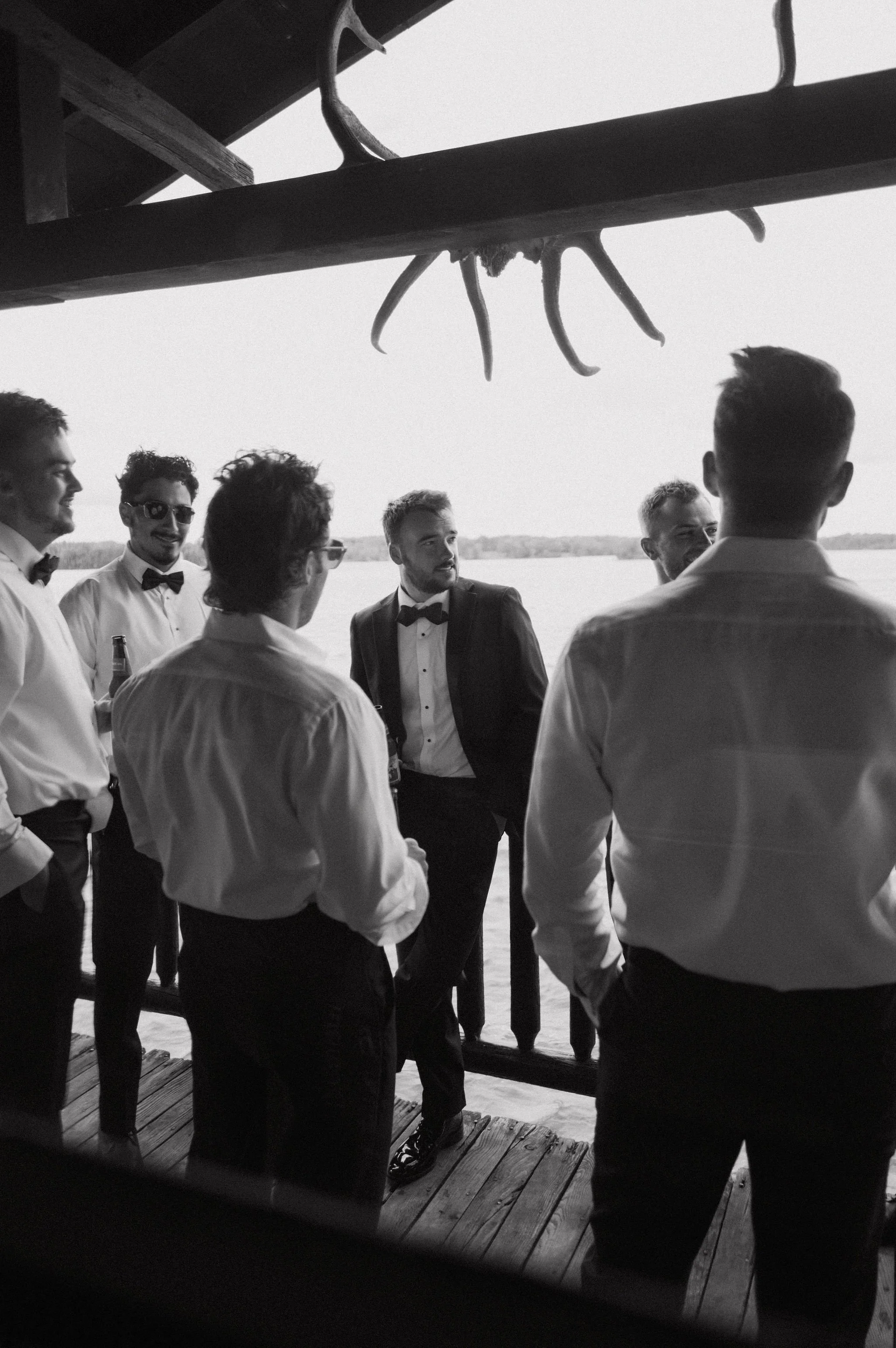 Group of men dressed in tuxedos and dress shirts on a deck by the water, with a large deer skull and antlers hanging overhead, black and white photo.