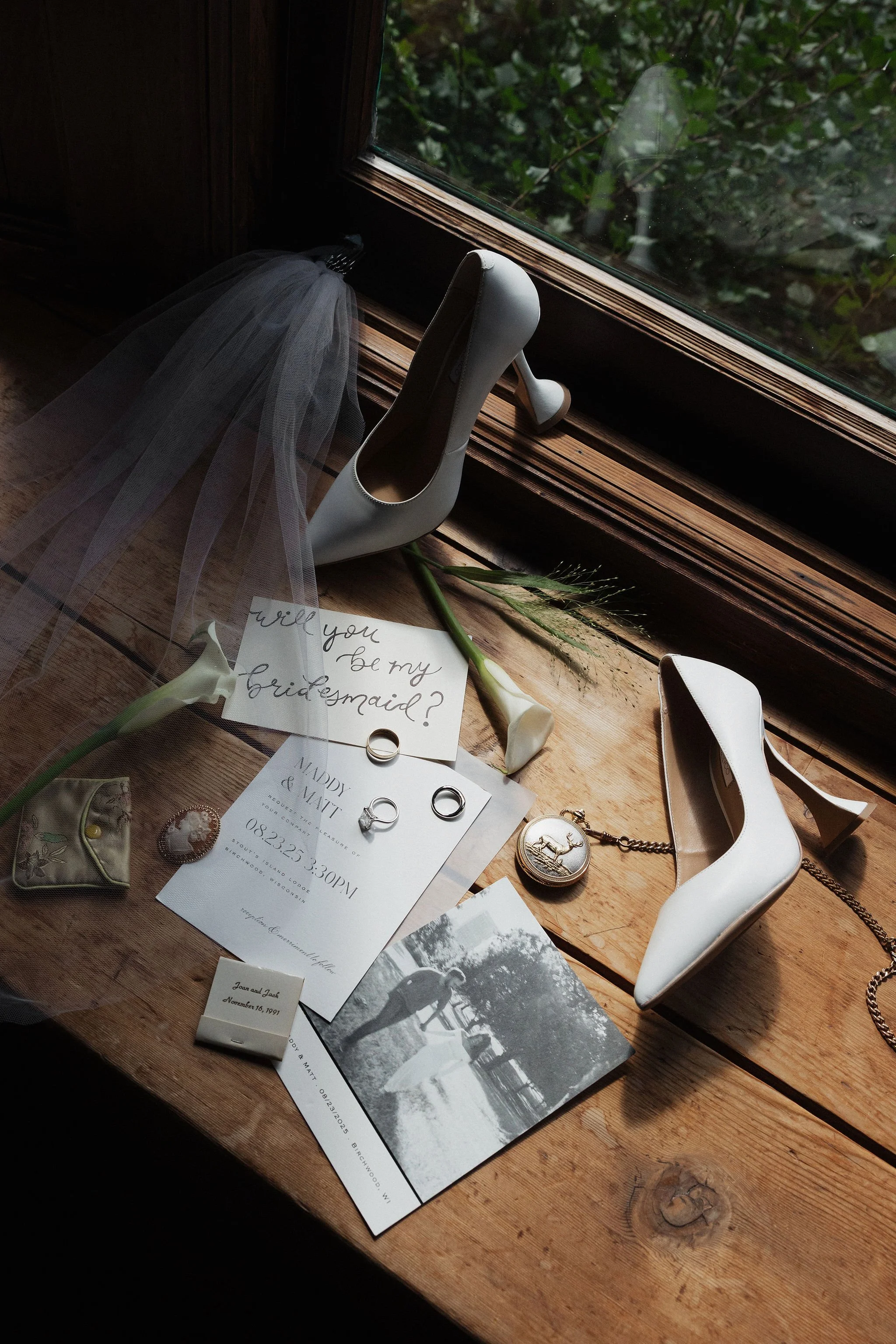 A wooden table near a window holding bridal accessories and wedding invitations, including a pair of white high heels, a veil, white calligraphy invitation, wedding rings, a vintage pocket watch, a bridal garter, a small box, a black-and-white photog