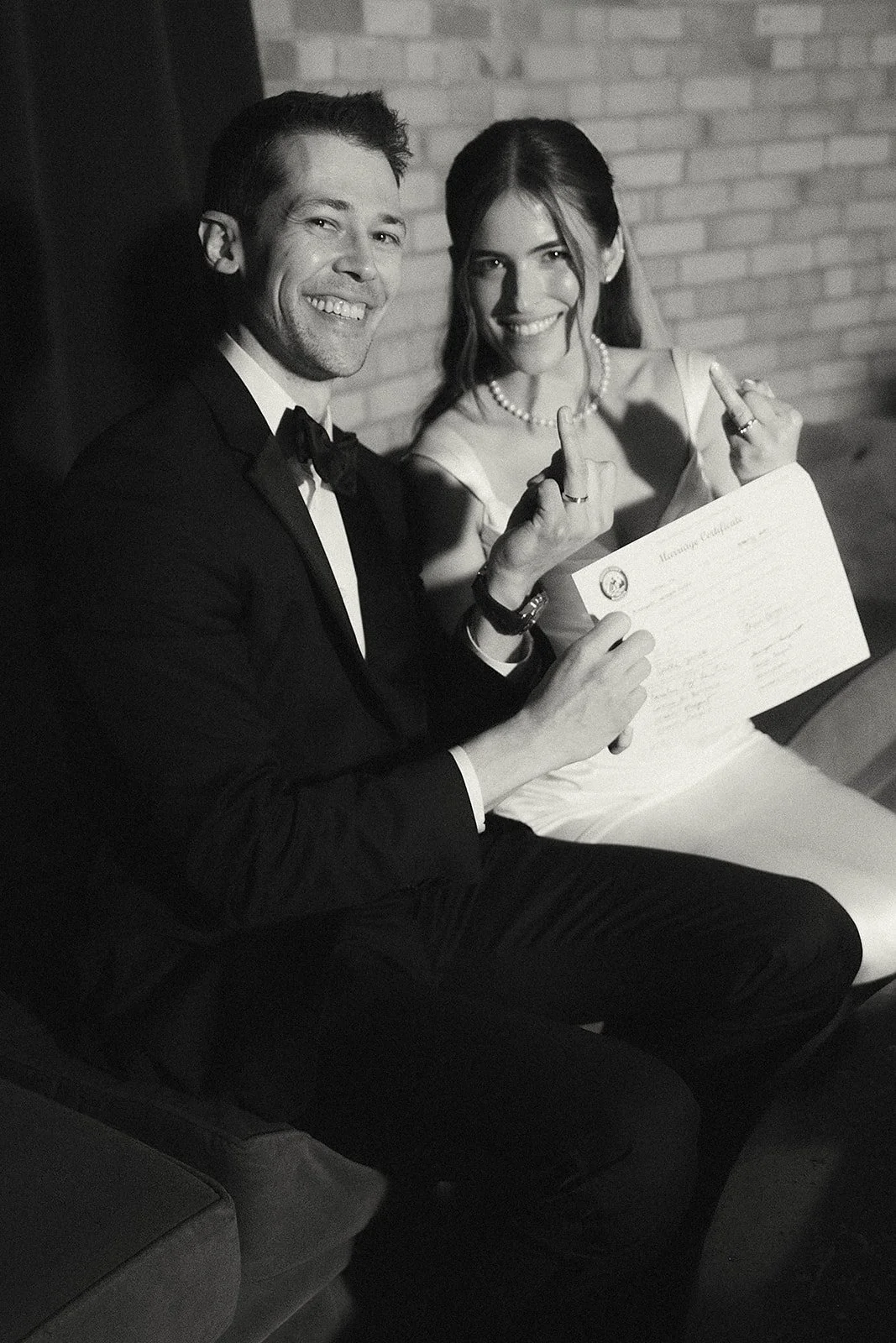 Black and white photo of a newlywed couple sitting on a couch, smiling, with the groom in a tuxedo and the bride in a white dress. They are holding a marriage certificate and both are showing middle fingers to the camera.
