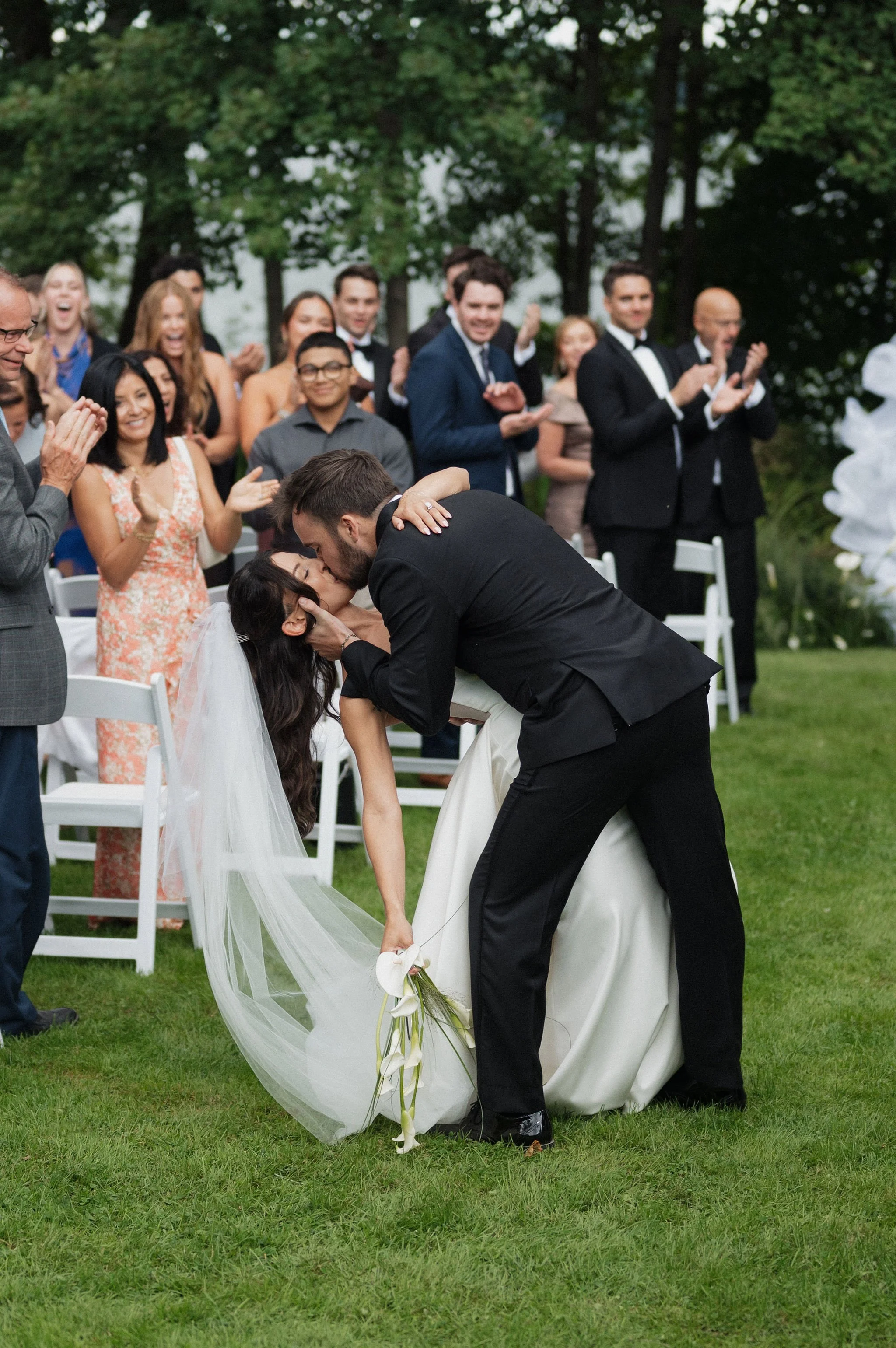 A bride and groom kissing during their outdoor wedding ceremony with guests clapping in the background.