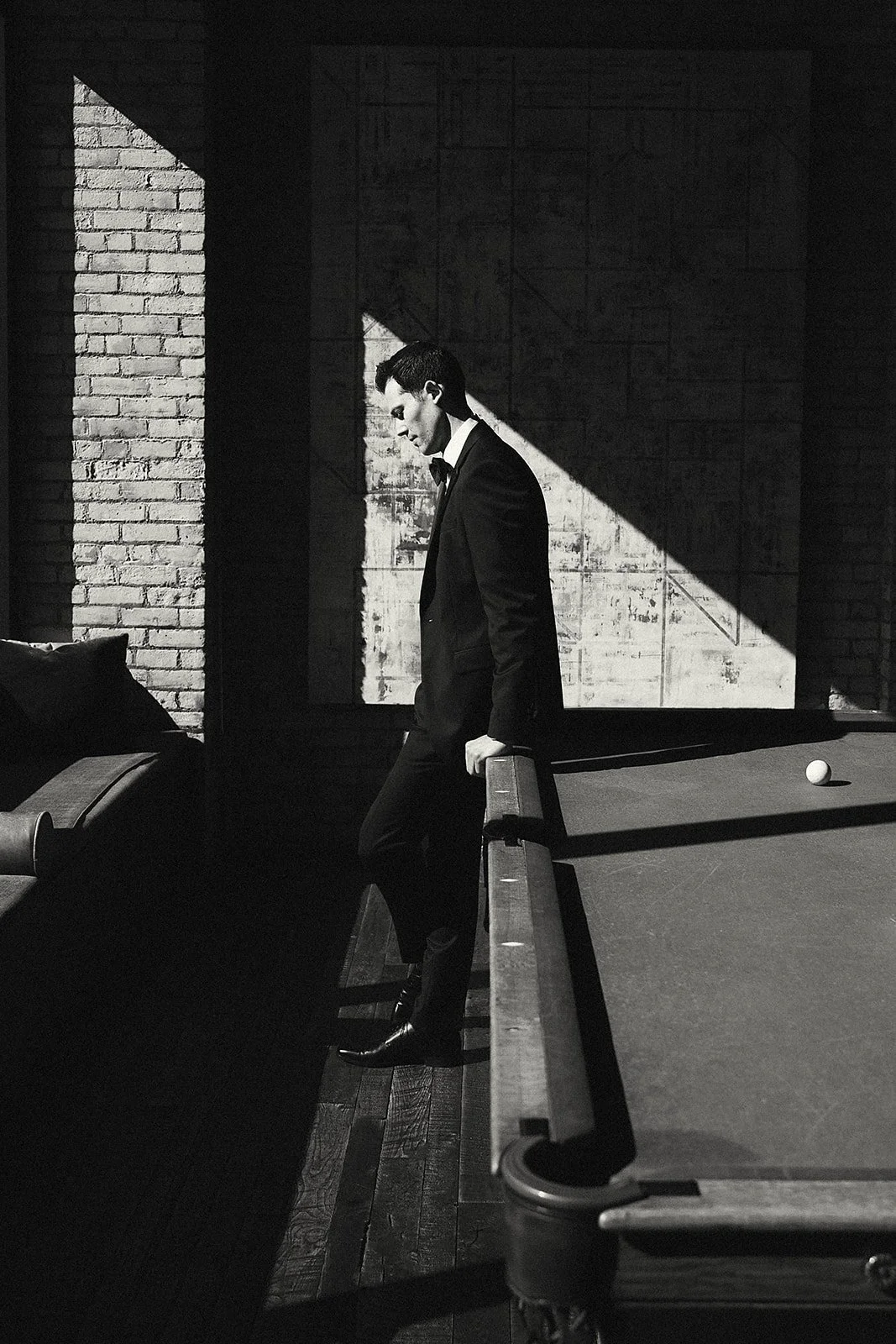 A man in a tuxedo on a pool table, holding a cue stick, in a dimly lit room with brick walls and shadows.