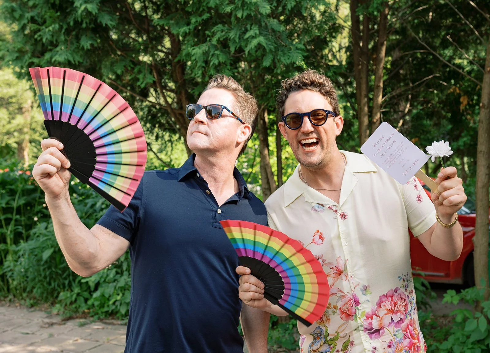 Two men holding rainbow-colored fans and celebrating outdoors, with trees in the background. One man is wearing a dark blue shirt and sunglasses, the other a floral shirt and glasses, holding a wedding invitation and a white flower.
