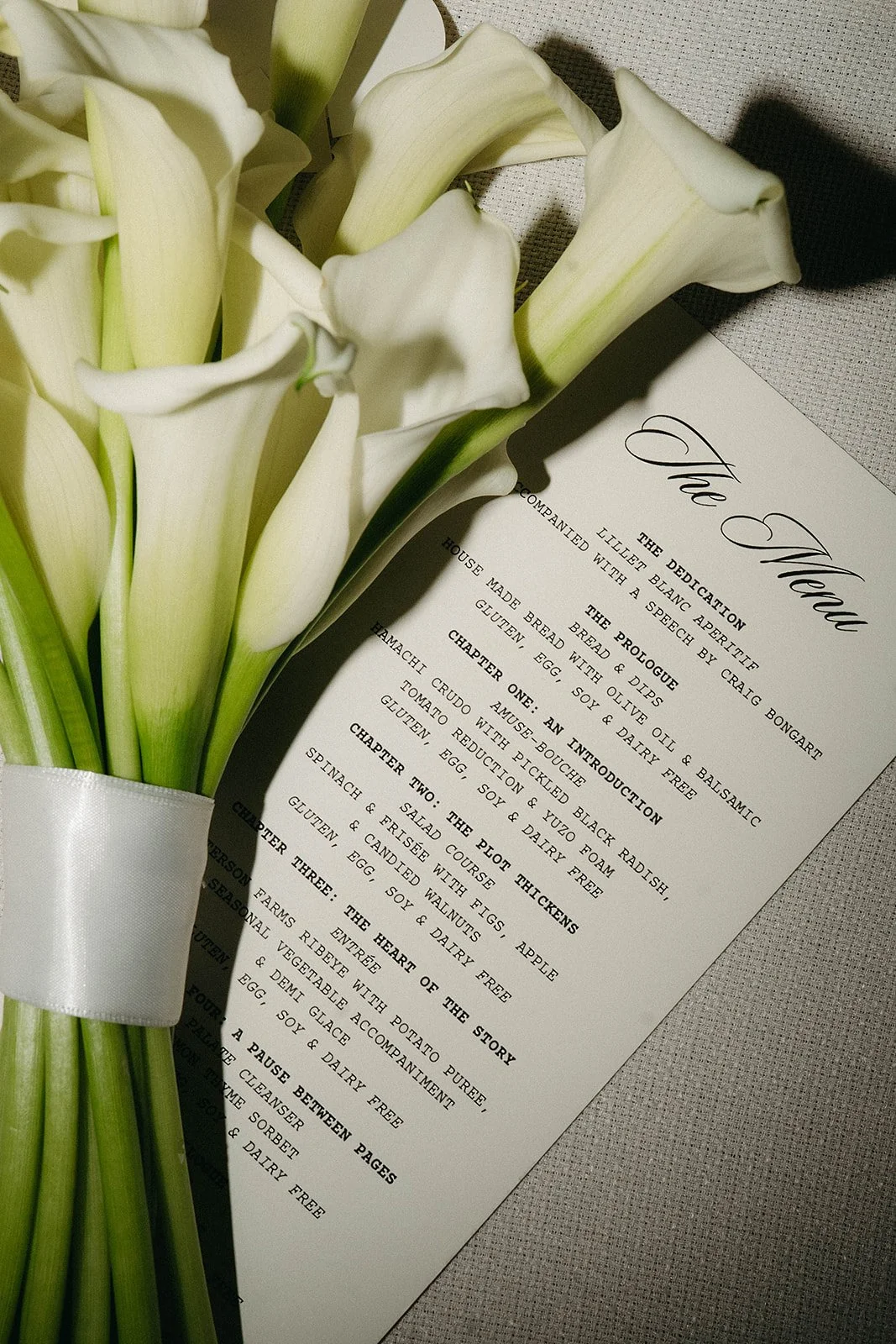 A white bouquet of calla lilies resting beside a printed menu on textured fabric.