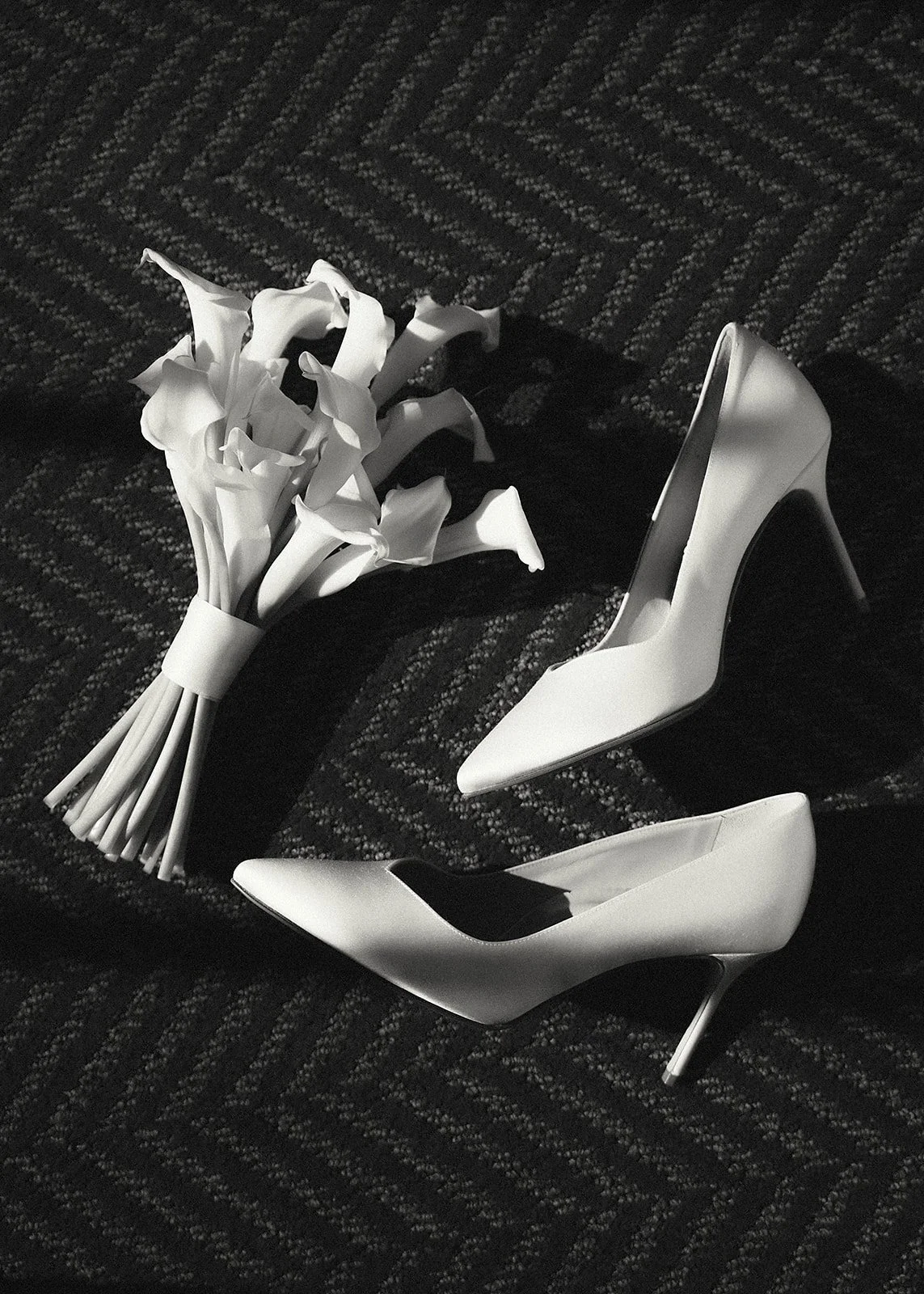 A pair of white high-heeled shoes and a small bouquet of white flowers placed on a dark textured surface.