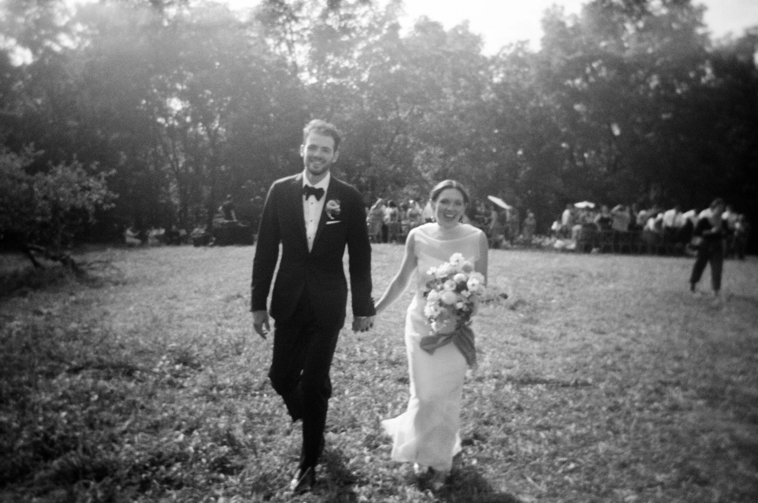 Black and white photo of a bride and groom walking hand in hand outdoors, with people and trees in the background.