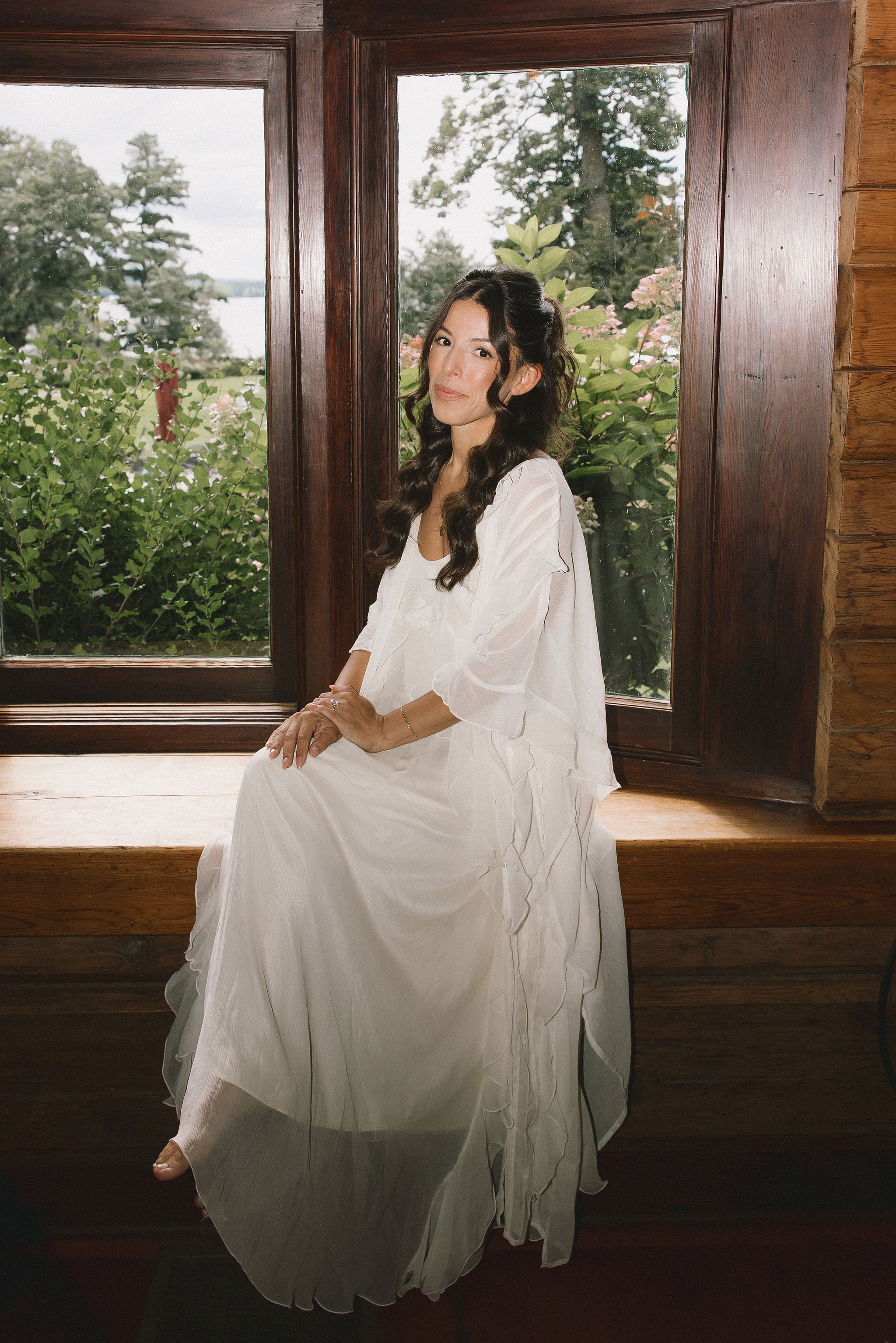 A woman with long wavy hair sitting on a wooden window seat inside a room, wearing a flowing white dress with ruffled sleeves, outdoors visible through the window.