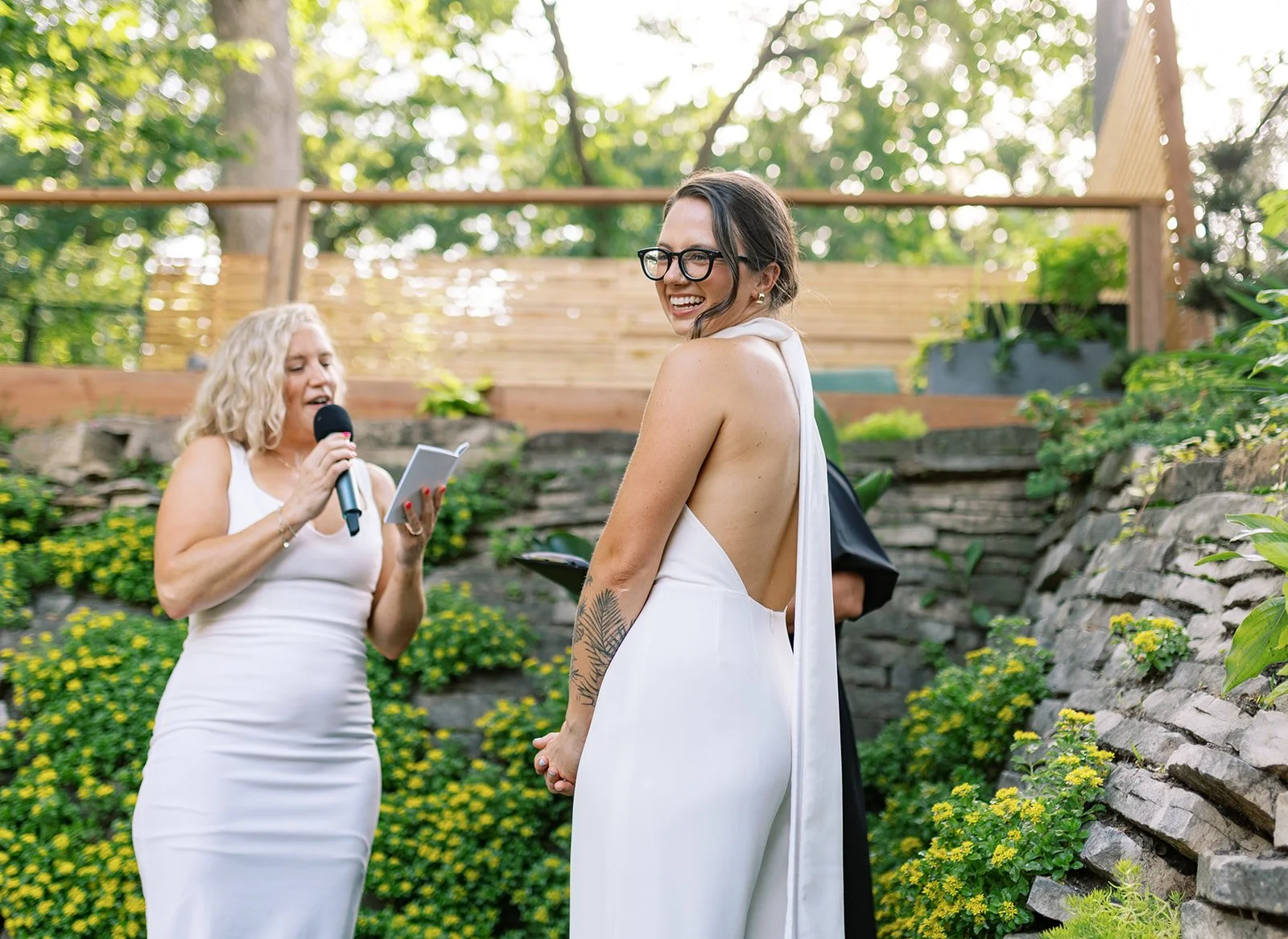 A woman in a white dress with an open back, holding hands with a woman in a sleeveless white dress, is smiling at a wedding ceremony outdoors in a garden surrounded by green plants and yellow flowers.