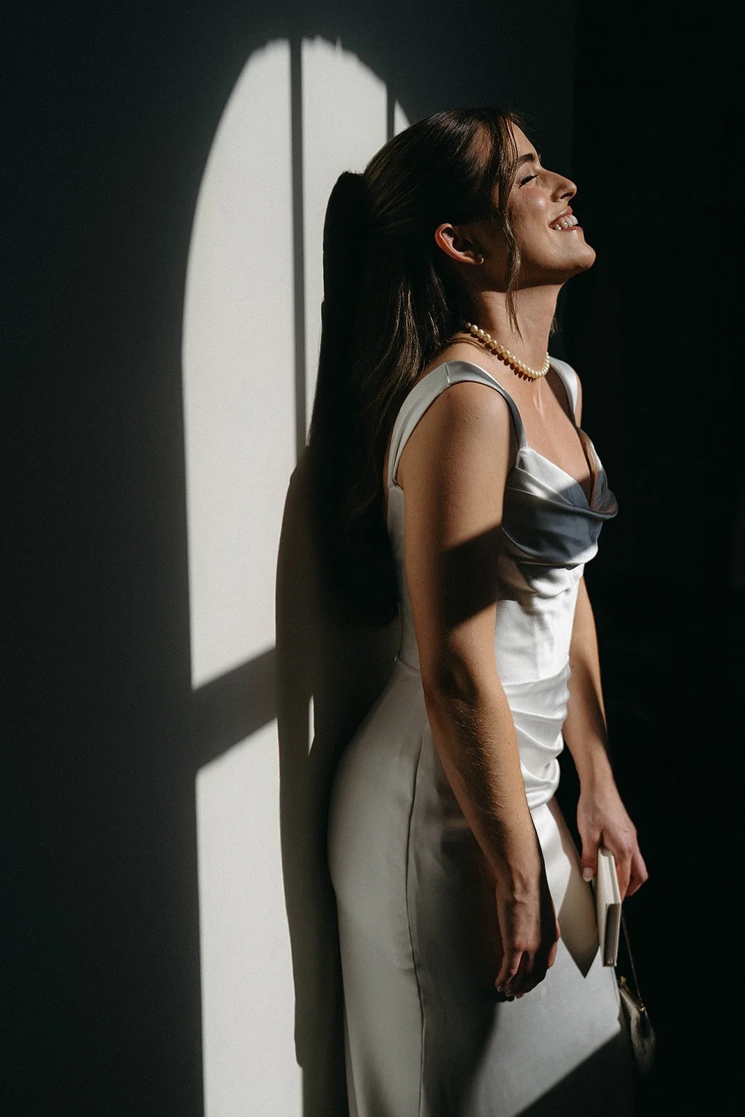 A woman in a silk dress, smiling with her eyes closed, standing against a wall with window-shadow patterns, holding a clutch and a phone.