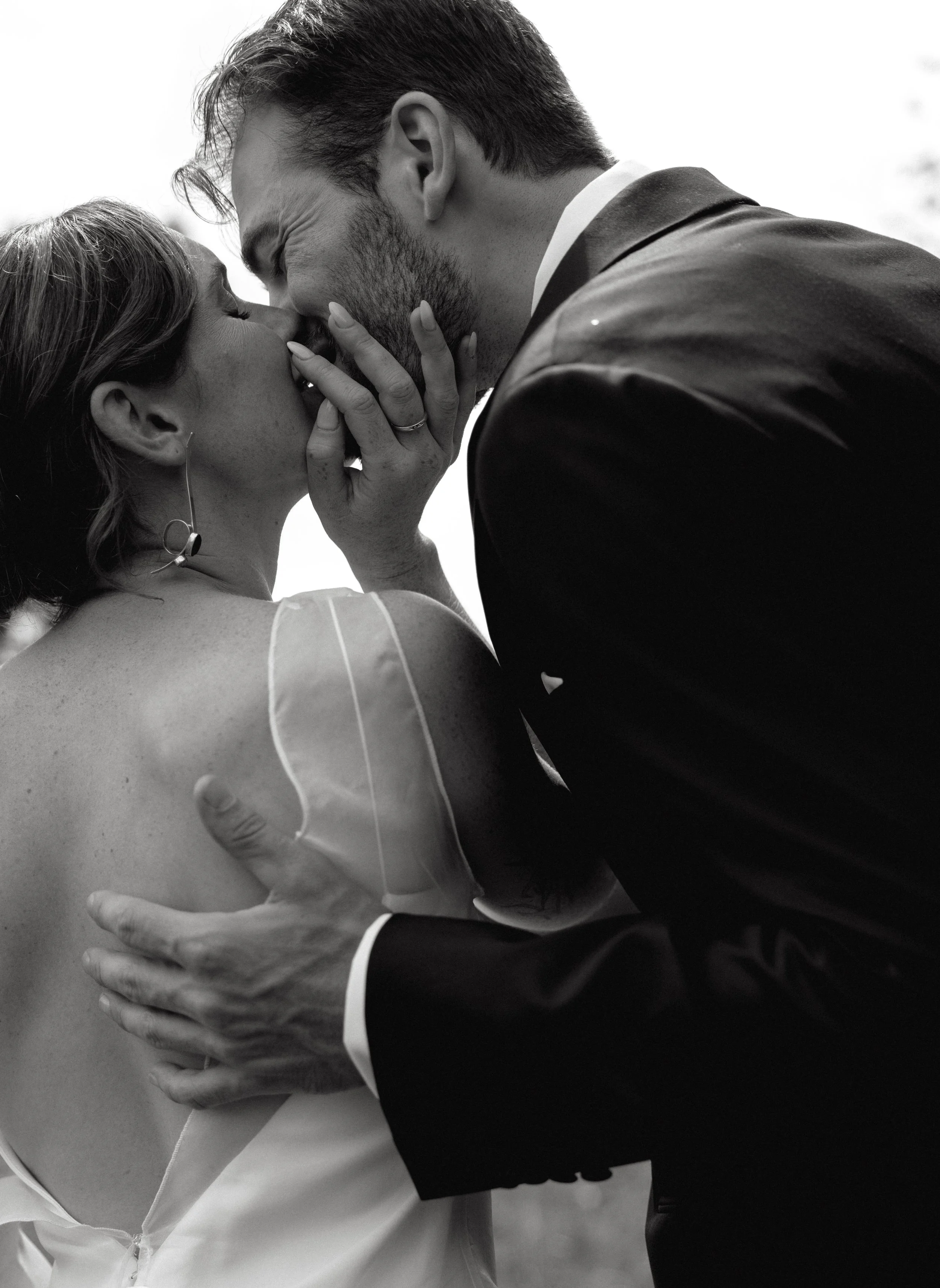 A black-and-white photo of a romantic moment between a bride and groom, kissing, with the bride touching the groom's face and the groom holding the bride's waist.