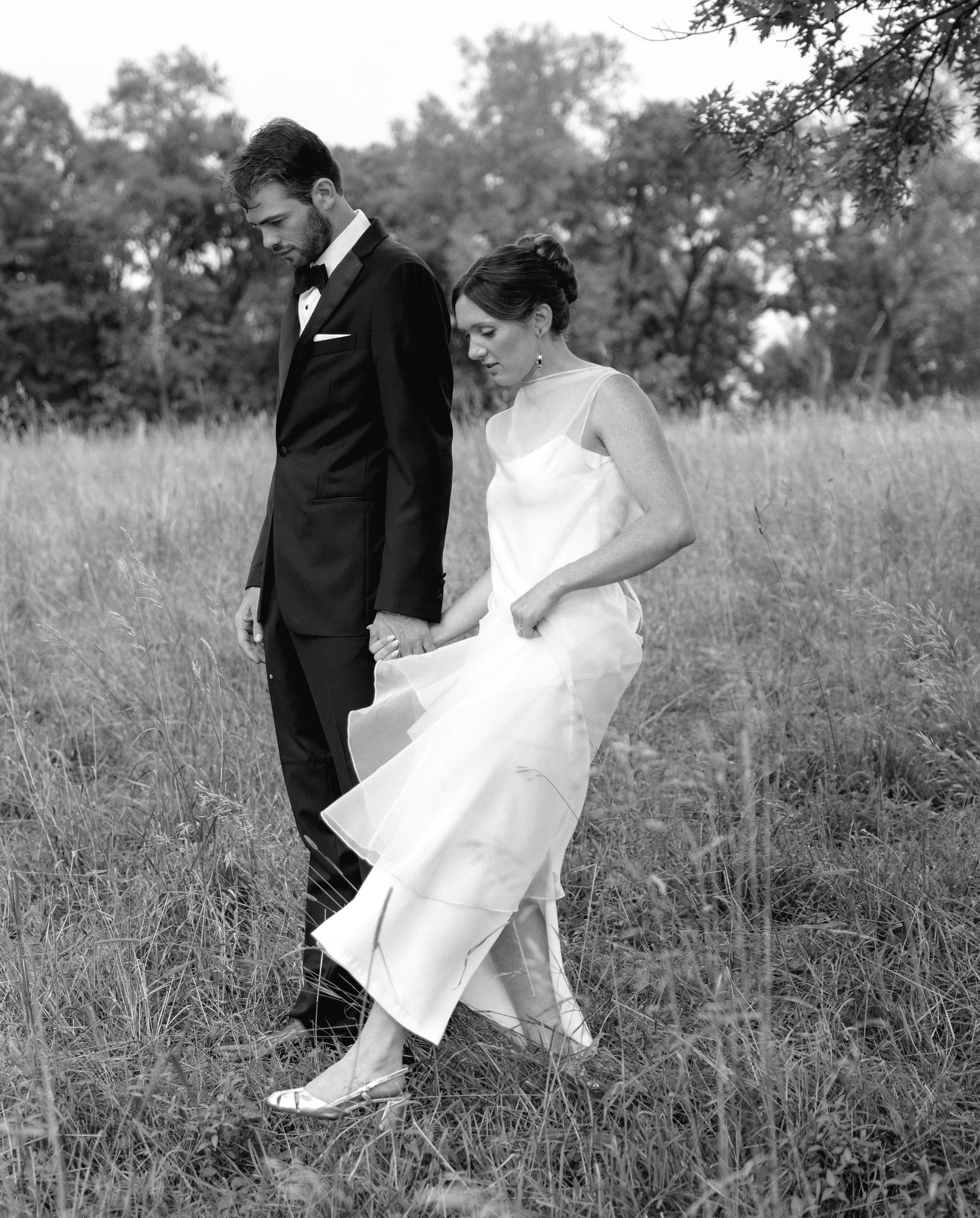 Black and white photo of a man and woman holding hands in a grassy field. The man is in a tuxedo, and the woman is in a white dress. They appear to be walking together, with trees in the background.