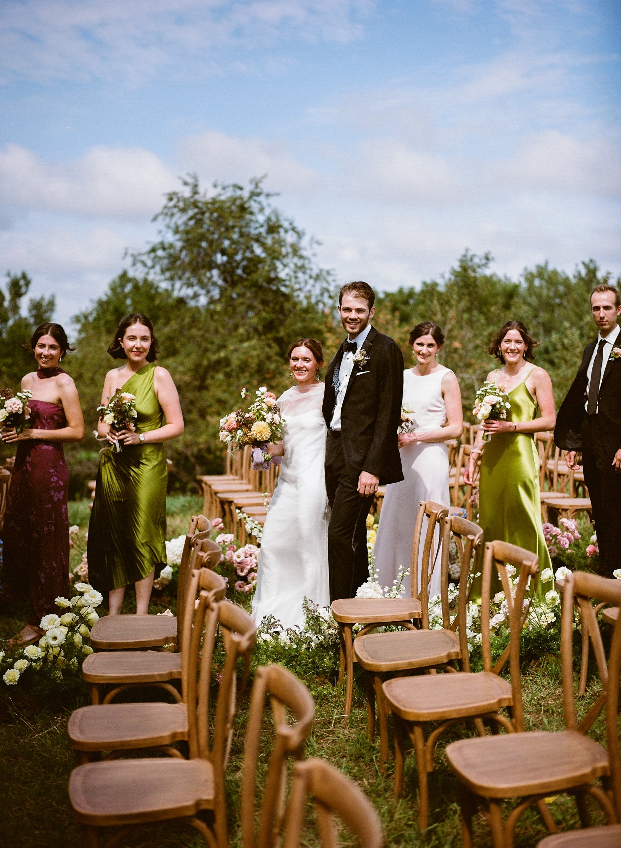 A bride and groom standing outdoors with bridesmaids and groomsmen during a wedding ceremony on a grassy field with chairs and floral decorations, under a blue sky with some clouds.