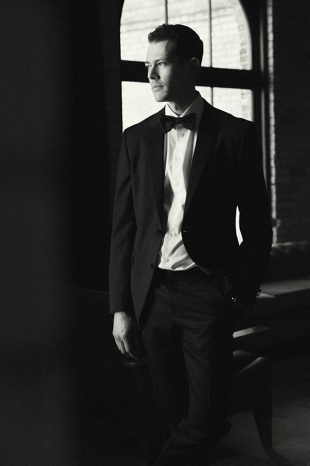 A man in a tuxedo with a bow tie, standing indoors near a window, looking out thoughtfully.