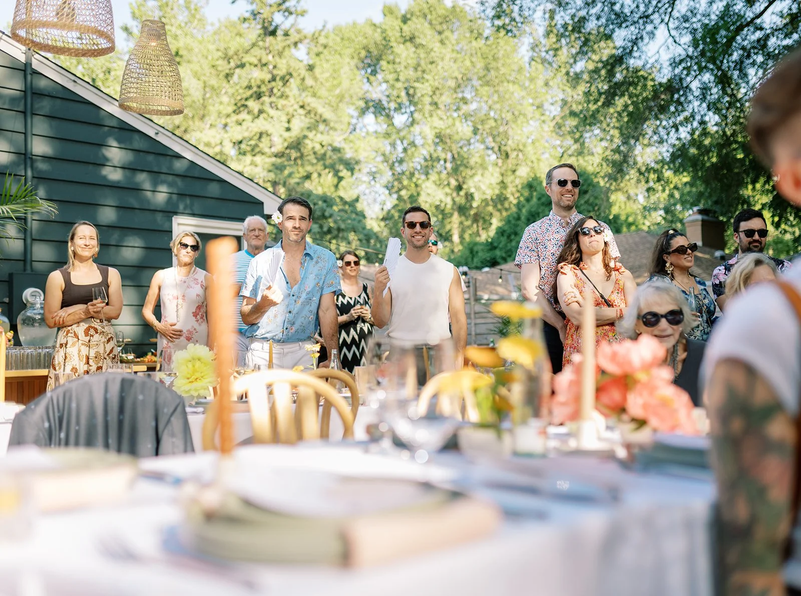 People gathered outdoors at a summer party or celebration, standing and smiling near a decorated table with flowers and candles, with trees and a house in the background.